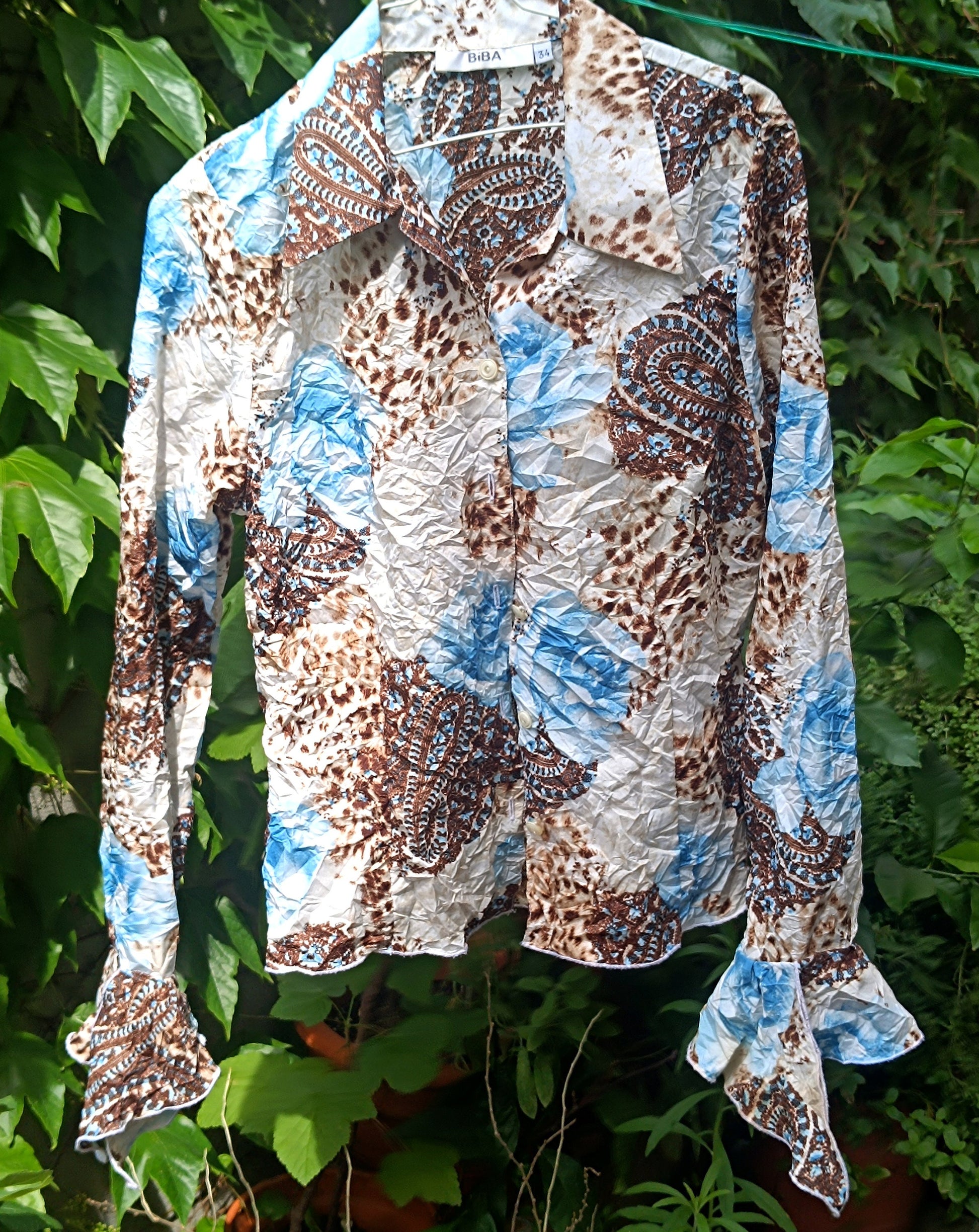 A vintage y2k floral print shirt with butterfly sleeves by the brand BiBA on a hanger, photographed against a lush green background of plants
