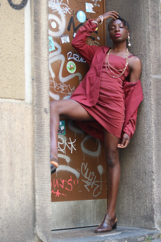 Stylish woman posing in urban setting, wearing a chic vintage red dress and jacket, accessorized with layered necklaces and earrings. Graffiti-covered door and weathered wall in the background add urban flair