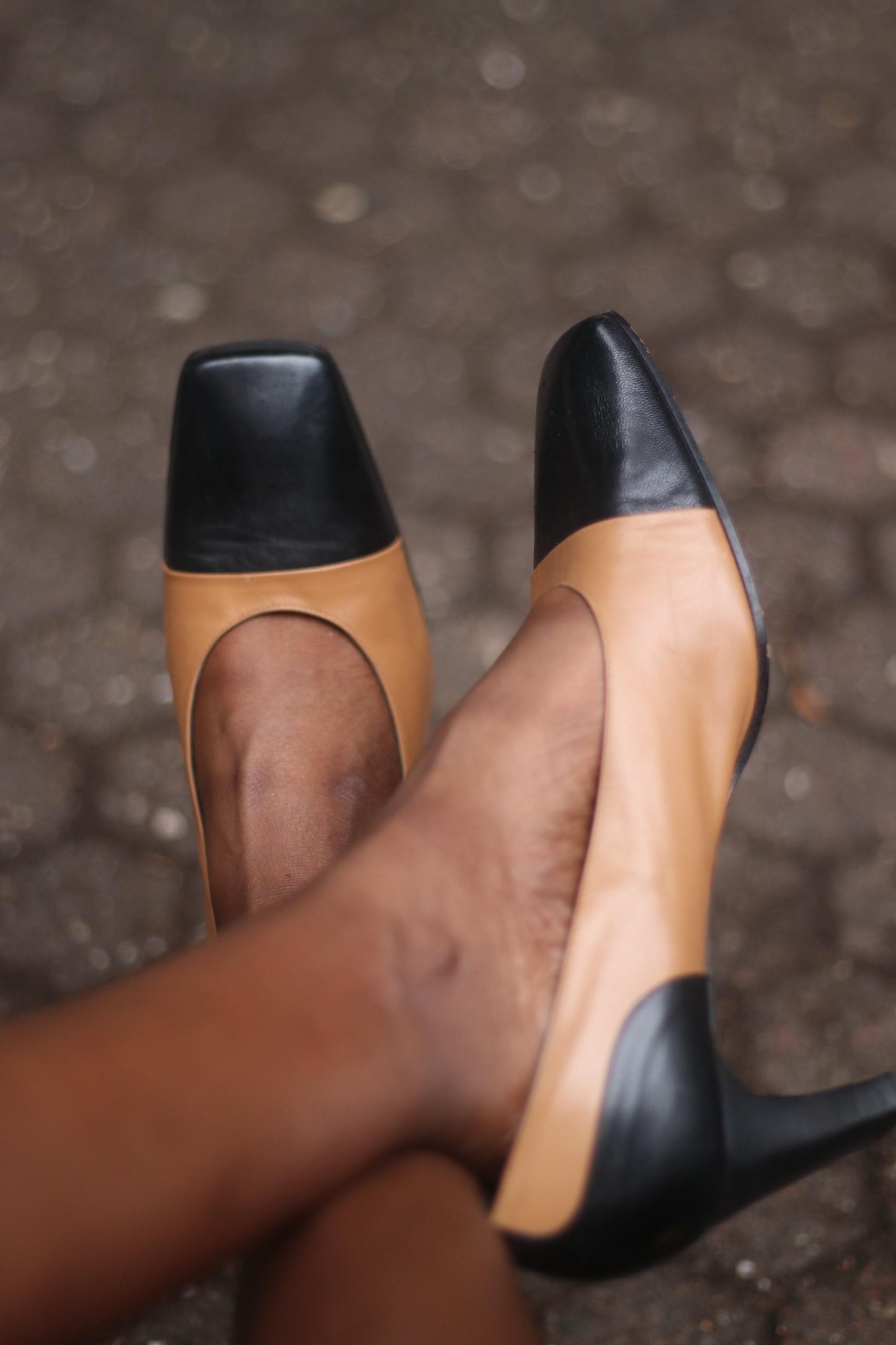 A pair of vintage two-toned high heels in brown and black on a model&#39;s feet. The brand is Tanja Jannetti. The shoes are photographed against a blurred background.