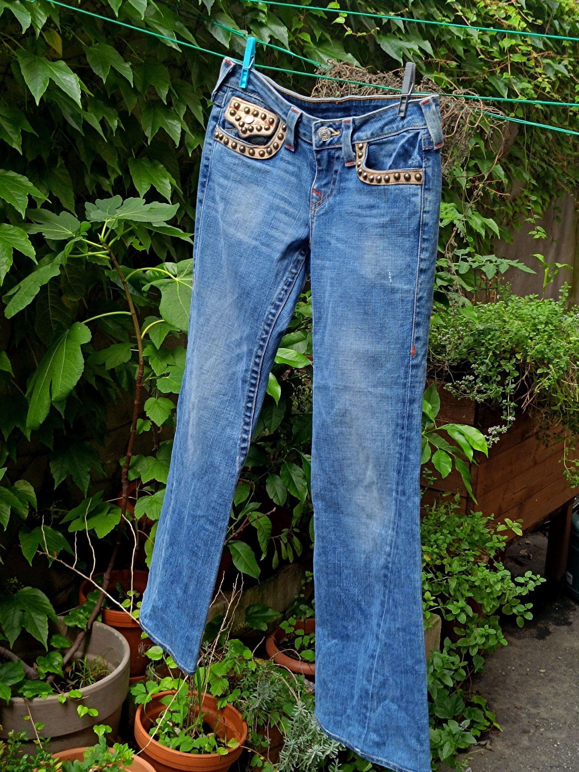 Light Blue &quot;True Religion&#39; jeans with studded metallic details on the front pocket is hung up with two clothing pegs on a laundry line. The designer y2k denim is photographed against a leafy green background.