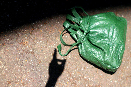 Vintage green bucket drawstring shoulderbag photographed lying on cobbled stone ground with a person&#39;s shadow visible nearby. The bag is leather and of high quality