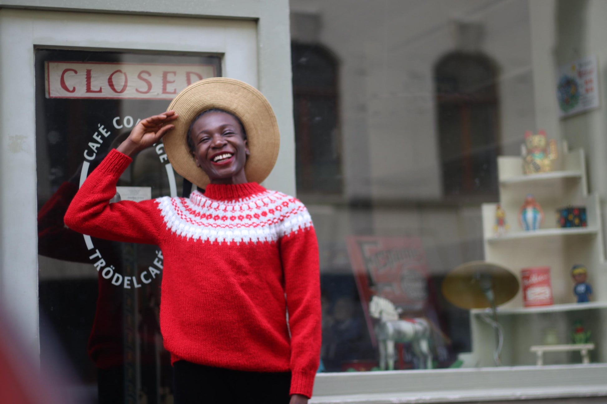 Black female model in a fashionable Scandinavian- inspired handmade red sweater with white patterns. She is wearing a vintage wide-brimmed hat smiling and posing happily in front of a shop window with a closed sign, enjoying a fun moment outdoors.
