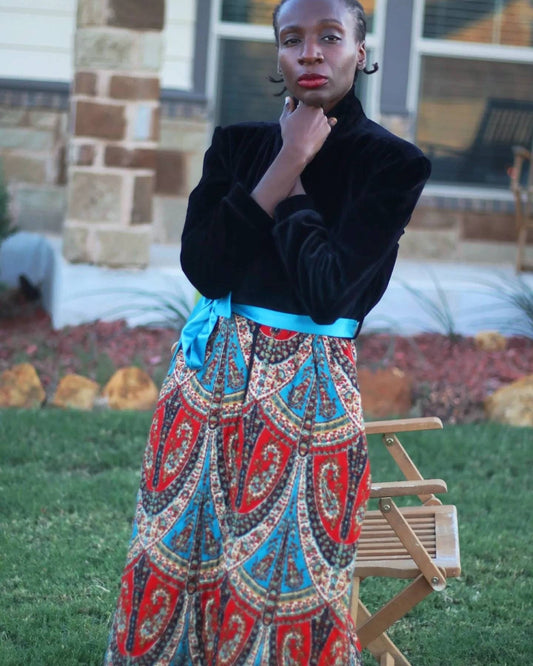 Model of African Heritage in a mix media maxi dress from the 80s. The unique vintage piece is black and velvety from top to waist and colorful, quilted and patterned till hemline. The dress also has a blue slash. She is standing in a garden