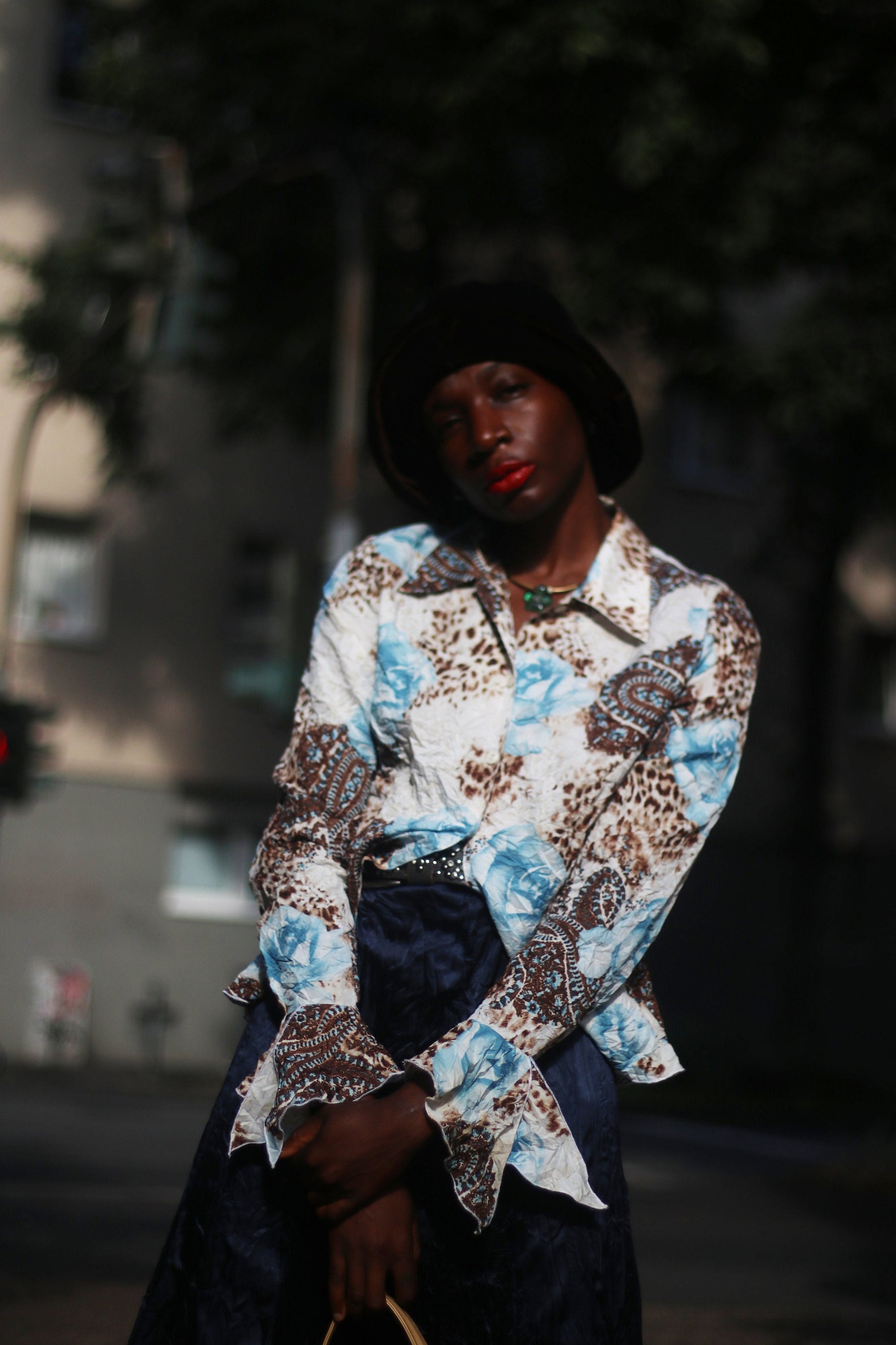 Model of African Heritage in a vintage y2k floral print shirt with butterfly sleeves and dark skirt, carrying a beige shoulder bag standing in sunlight.