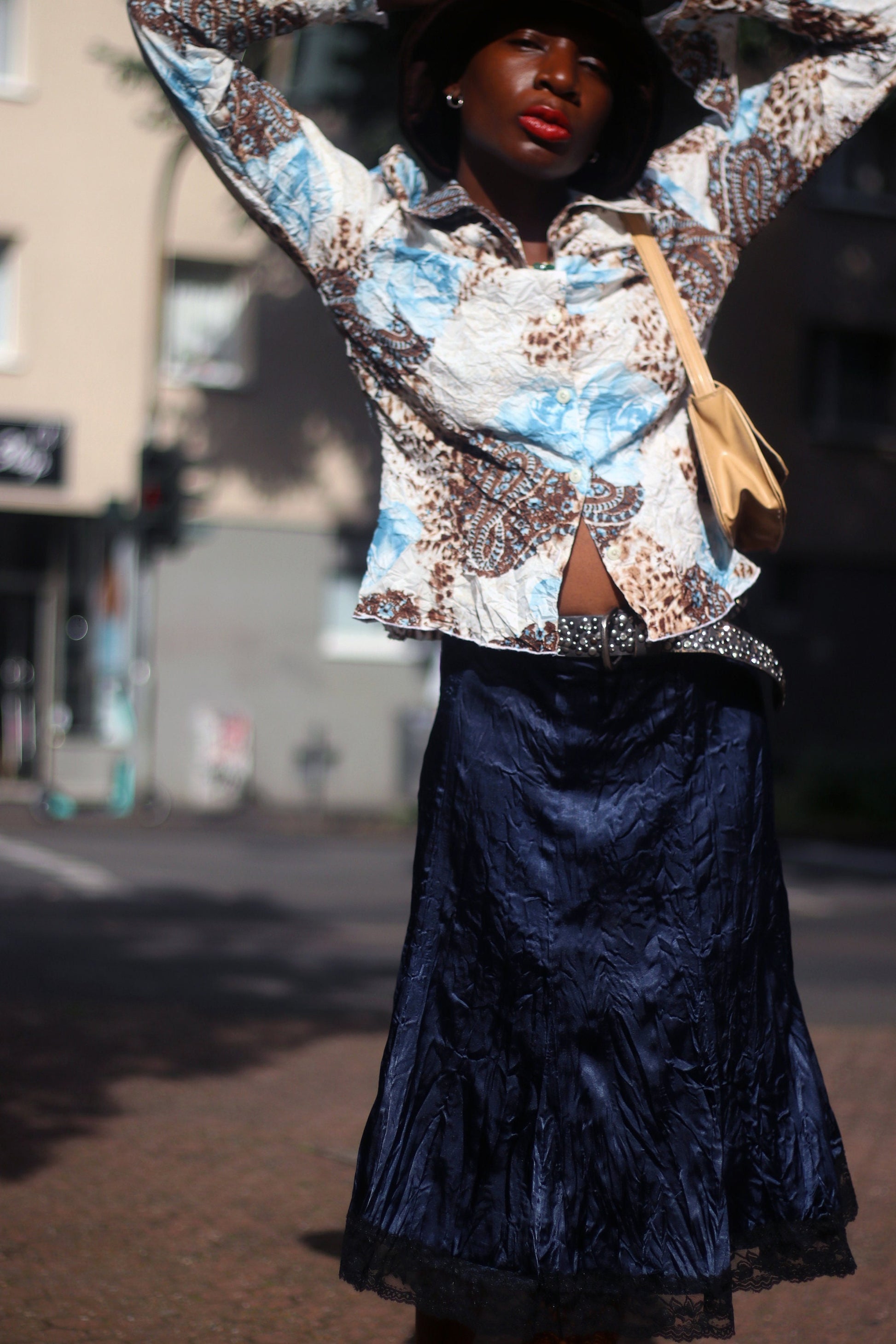 Model of African Heritage in a vintage y2k floral print shirt with butterfly sleeves and dark skirt, carrying a beige shoulder bag standing in sunlight.