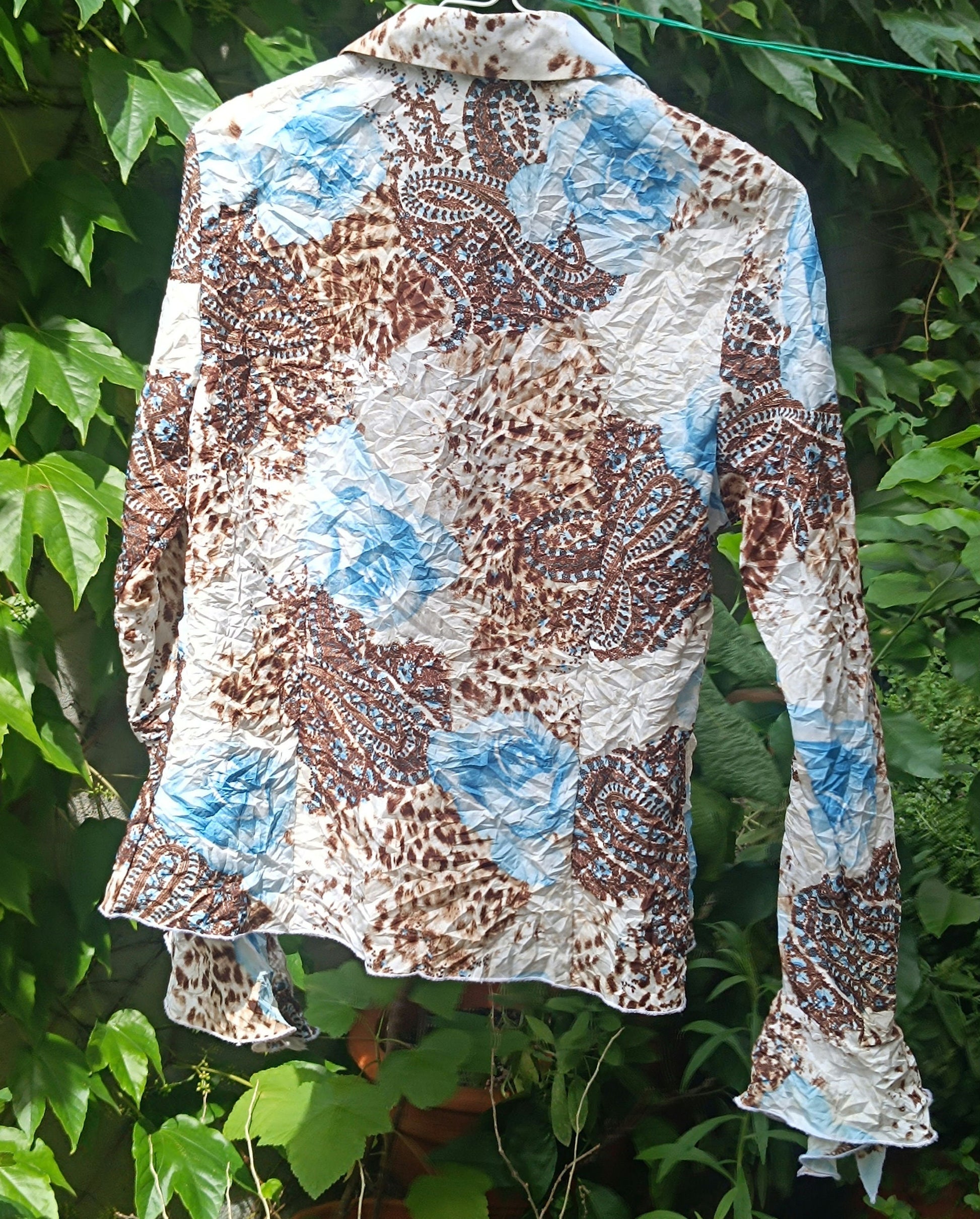 A vintage y2k floral print shirt with butterfly sleeves by the brand BiBA on a hanger photographed against a lush green background of plants