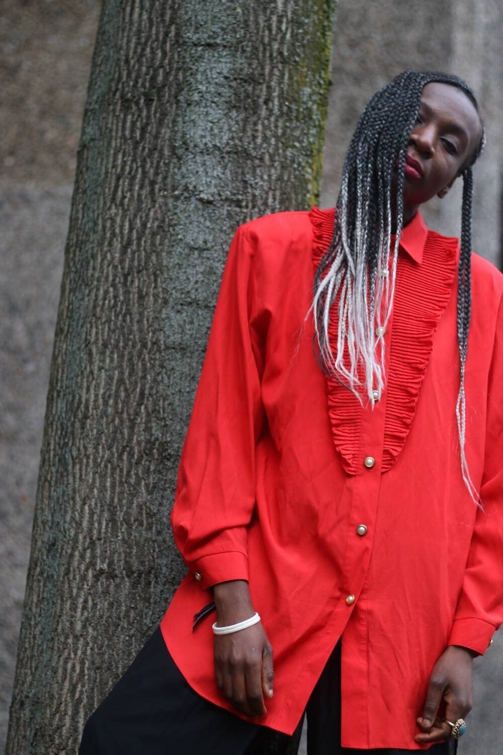 Fashionable model of African heritage wearing a vintage vibrant red button-up shirt with ruffle detailing, complemented by long, braided hair with silver accents, poses elegantly against a tree trunk.