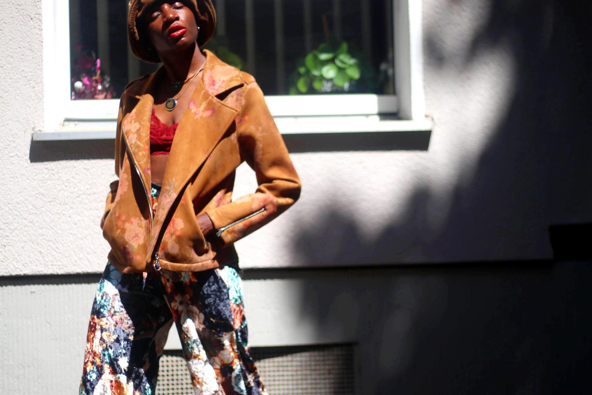 A woman of African heritage wearing a vintage Y2K floral jacket and maximalist floral pants is photographed infront of an urban window. The Boho outfit is complemented with a hat, a red cropped lace top and two necklaces. The jacket is Italian made.
