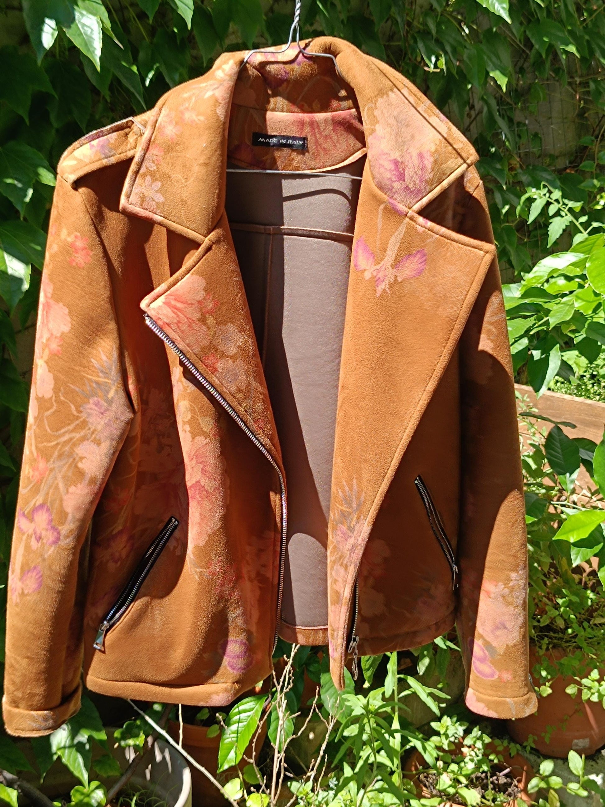 A vintage Y2K floral jacket on a hanger is photographed against a background of green plants. The brown jacket is open and has two zipped side pockets.