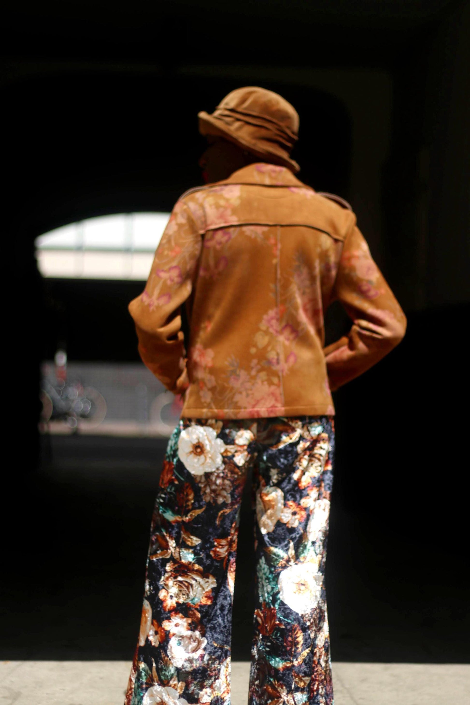 A woman of African heritage wearing a vintage Y2K floral jacket and maximalist floral pants is photographed to in the sun. Her back is to the camera. The outfit is complemented with a hat, a red lace top and two necklaces. The jacket is Italian made.