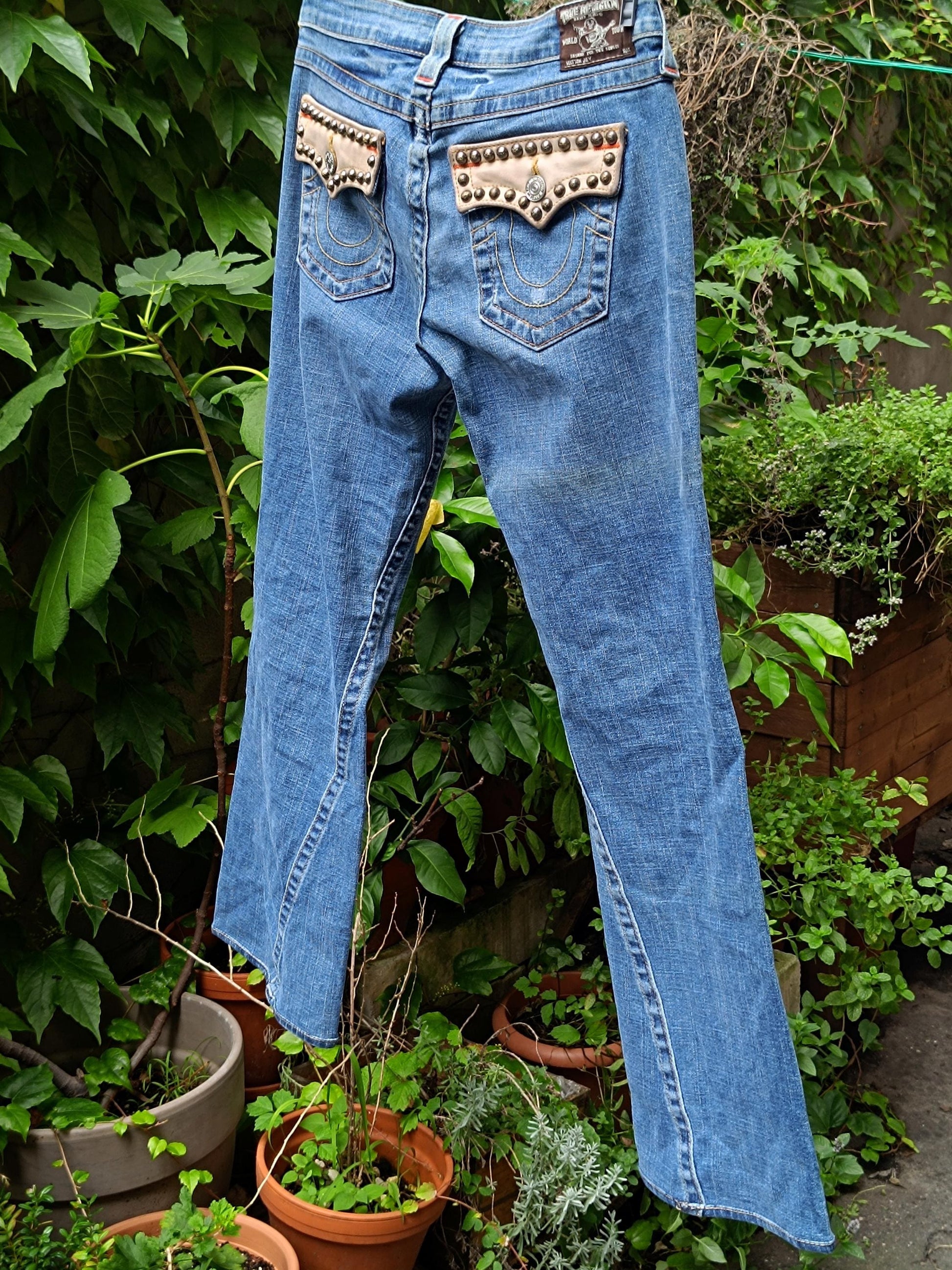 A pair of women&#39;s &quot;True Religion&#39; jeans with studded back pocket details is photographed against a leafy green background. The y2k designer denim is light blue and has a flared hemline