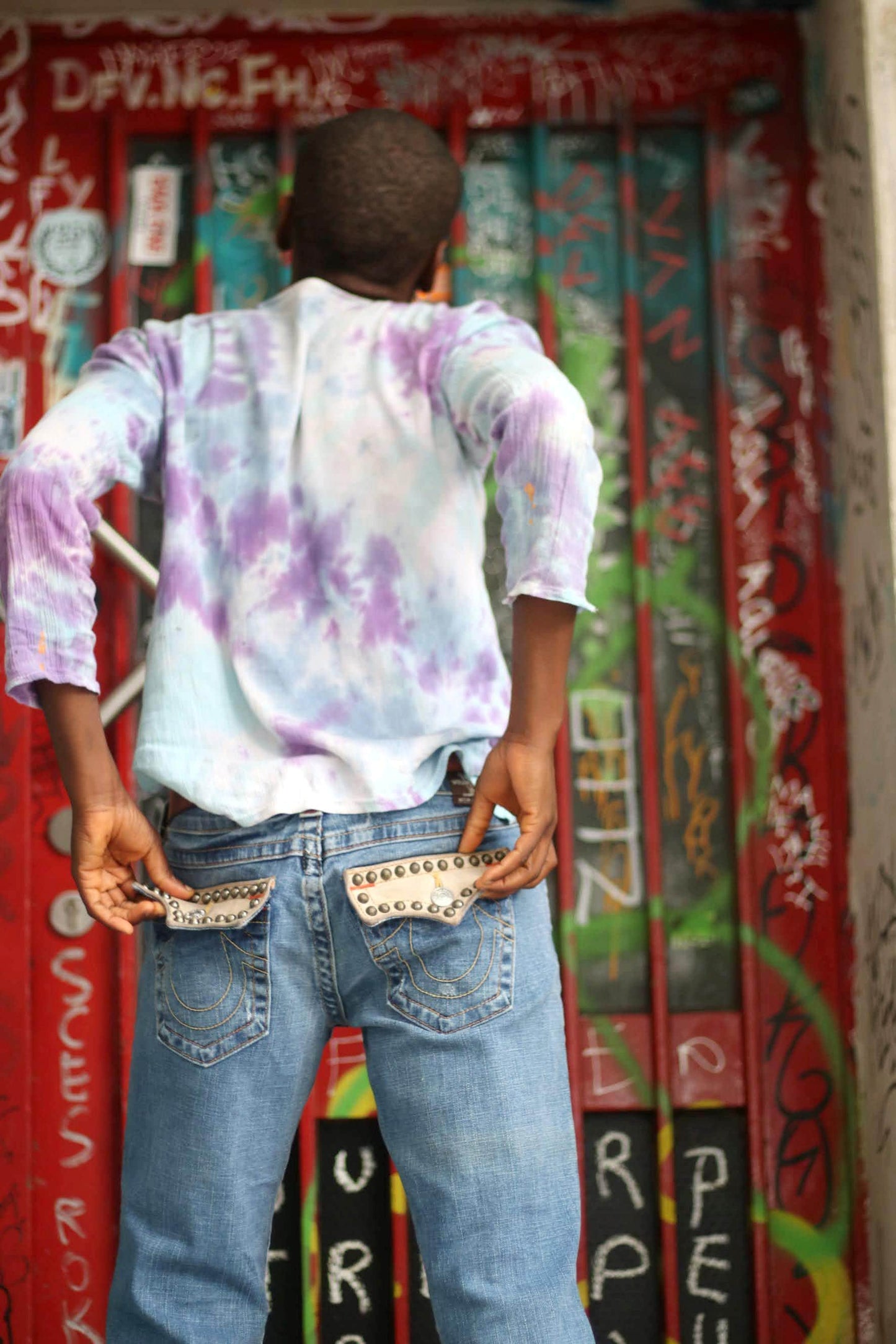A black bald woman with her back to the camera in pair of &quot;True Religion&quot; jeans and a vibrant tie-dye shirt . She stands against a graffiti covered door and the studded details on the back pockets of the designer y2k blue jeans are showcased.