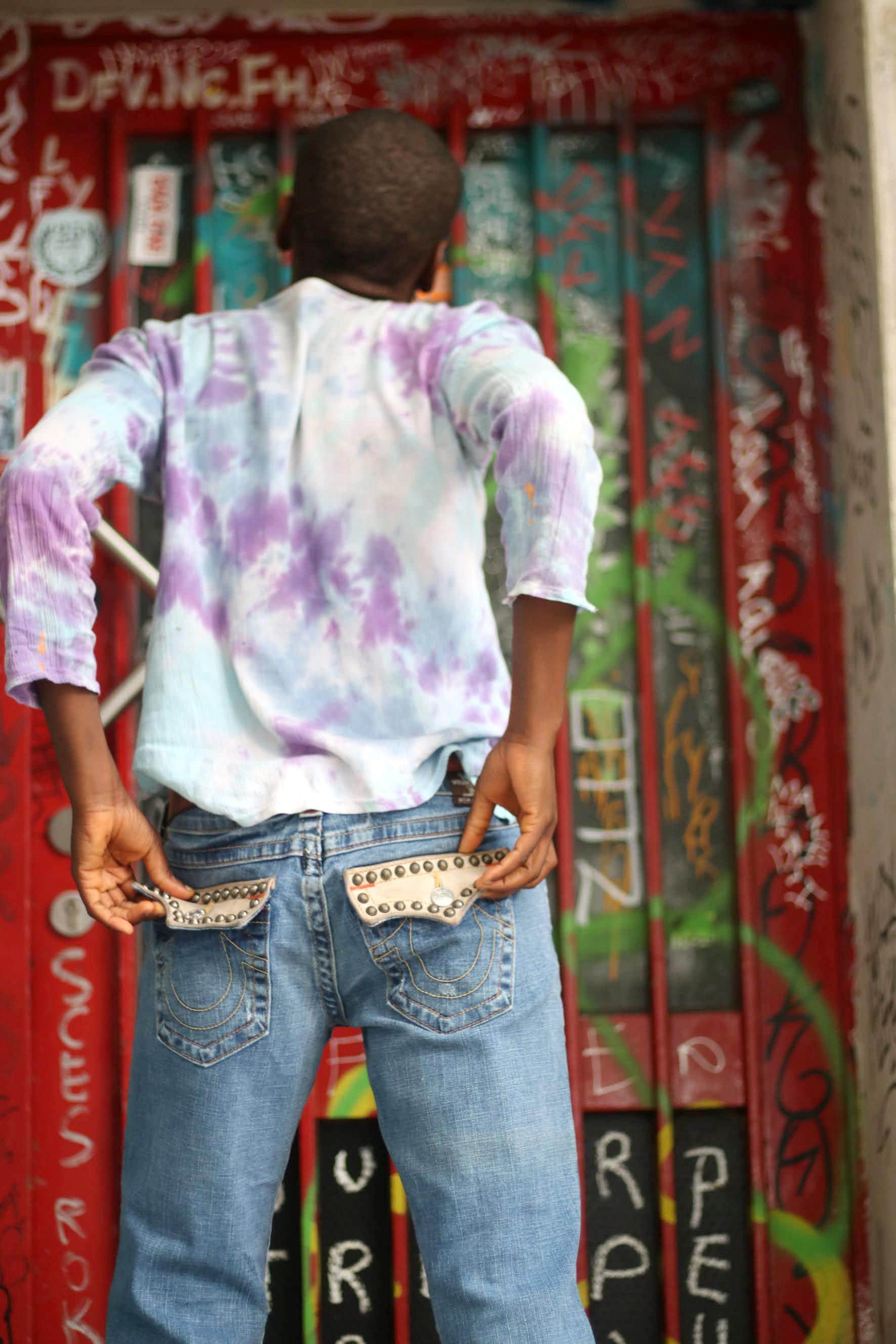 A black bald woman with her back to the camera in pair of &quot;True Religion&quot; jeans and a vibrant tie-dye shirt . She stands against a graffiti covered door and the studded details on the back pockets of the designer y2k blue jeans are showcased.
