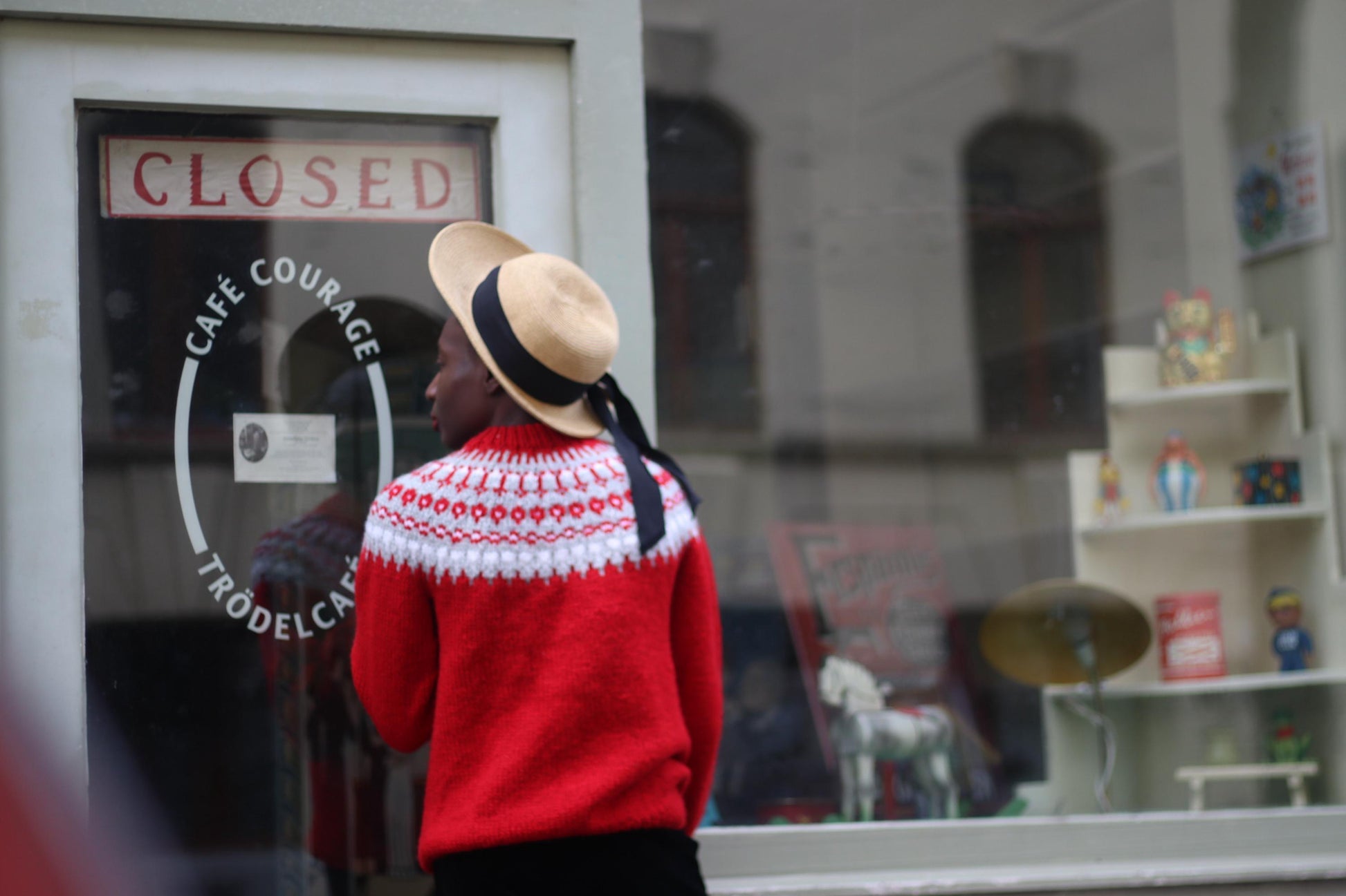 Fashionable model in a vintage Scandinavian red sweater with white patterns, black trousers and wide-brimmed hat. She is in front of a shop window with a closed sign, her back to the camera , looking into the shop