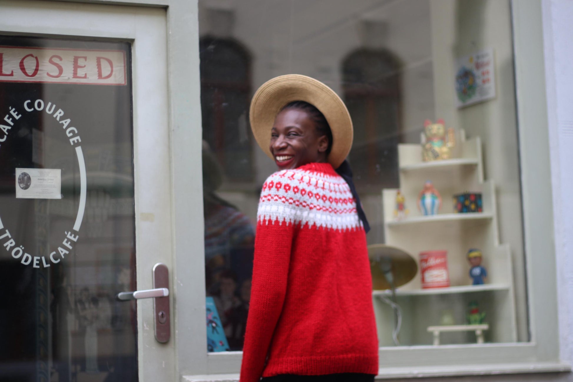 Fashionable black model in a trendy vintage Scandinavian red sweater with white patterns by the neckline, black trousers and wide-brimmed hat. She is in front of a shop window with a closed sign, smiling and looking over her shoulder