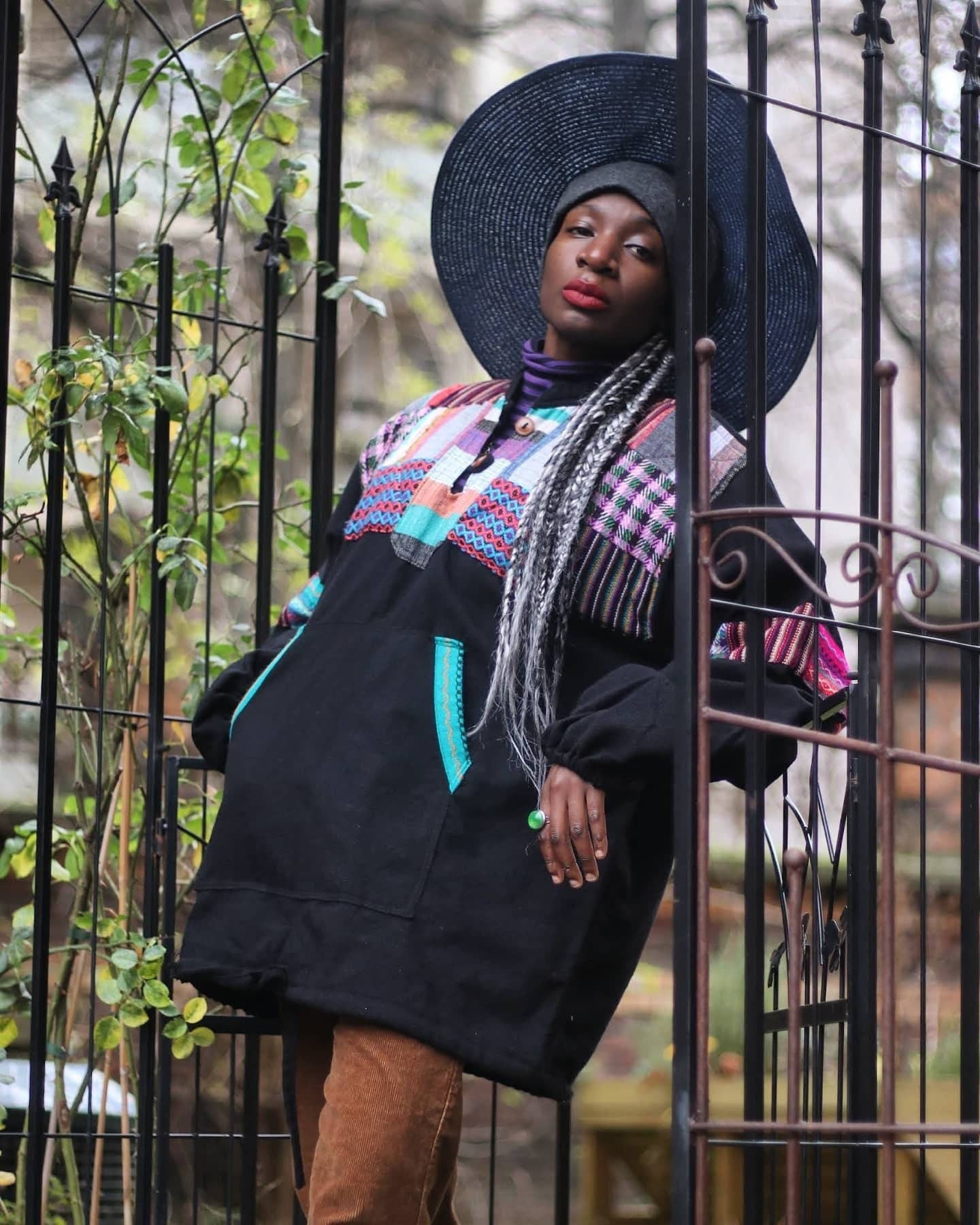 Female model in a vintage black Ecuadorian patchwork sweater photographed standing in a lush green background. She wears a purple striped turtleneck underneath the ethno chic long sleeves design , a blue hat and brown corduroy pants.