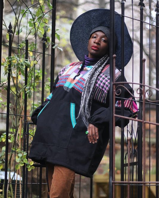 Female model in a vintage black Ecuadorian patchwork sweater photographed standing in a lush green background. She wears a purple striped turtleneck underneath the ethno chic long sleeves design , a blue hat and brown corduroy pants.