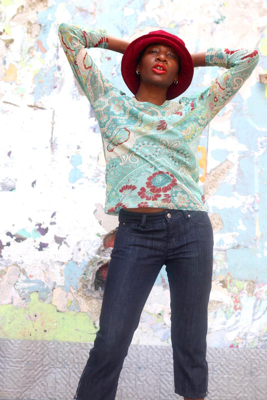 Model in a y2k floral psychedelic turquoise top paired with denim capris.She hason a red hat and is photographed against an arty collage-like call in an urban setting.
