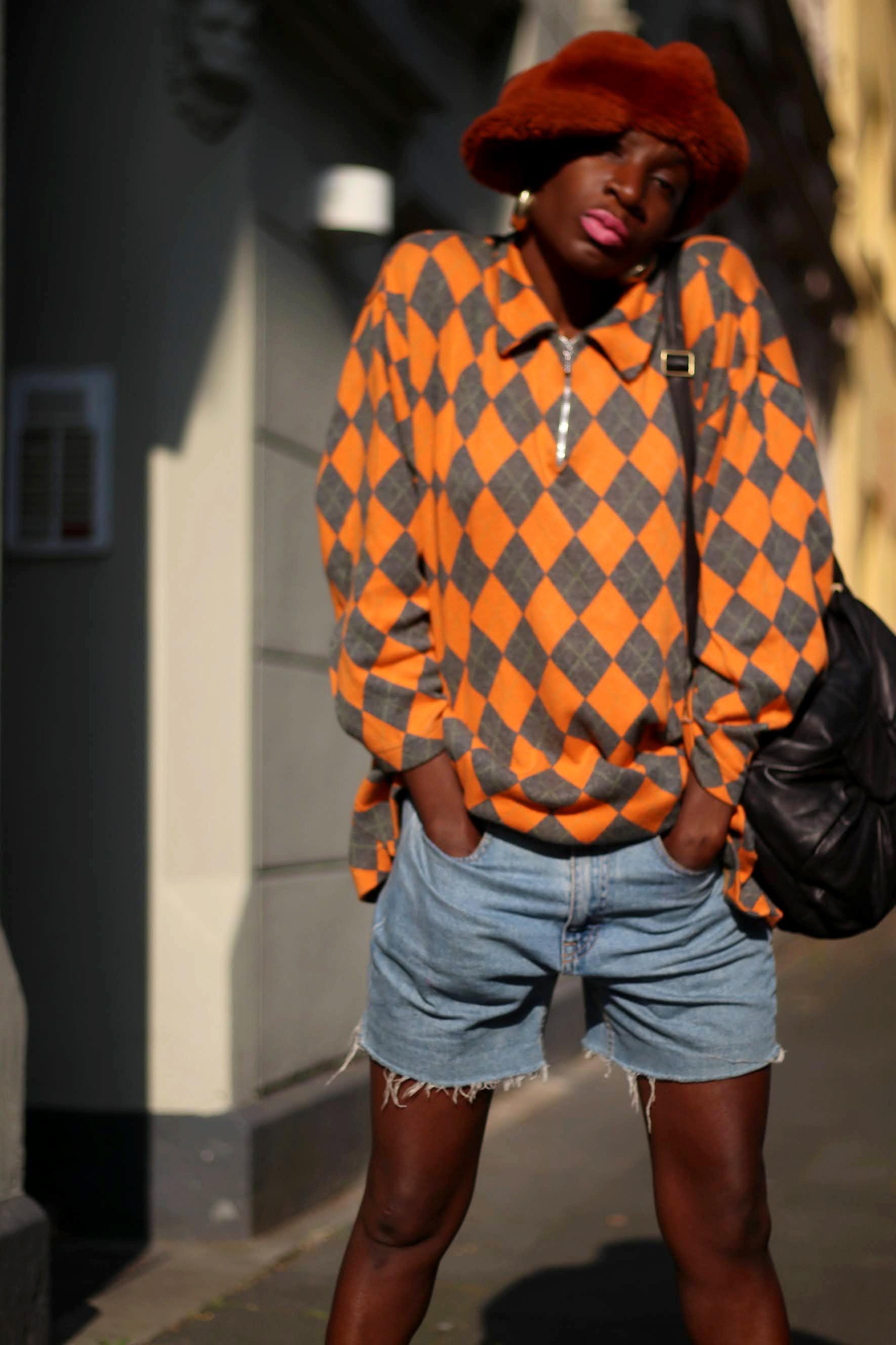 Female model in a vibrant orange and grey checkered sweater paired with denim shorts and a fashionable hat, photographed on the streets. She carries a black bag and both hands are in her front pocket