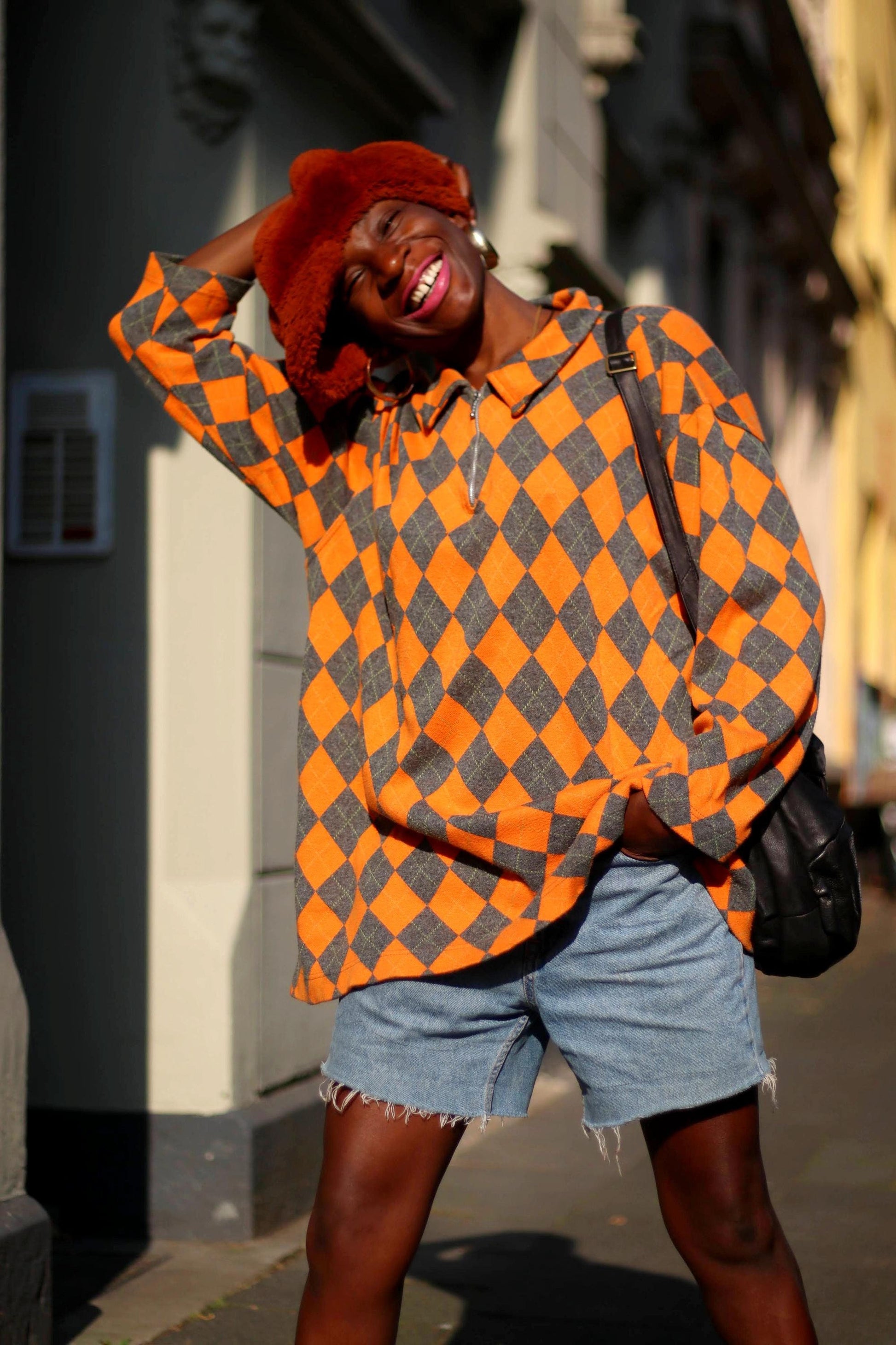 Stylish individual in a vibrant vintage orange and grey checkered sweater paired with denim shorts and a fashionable hat, smiling warmly outdoors. Expert in street fashion, casual outfit inspiration, sunny day attire, and urban style.