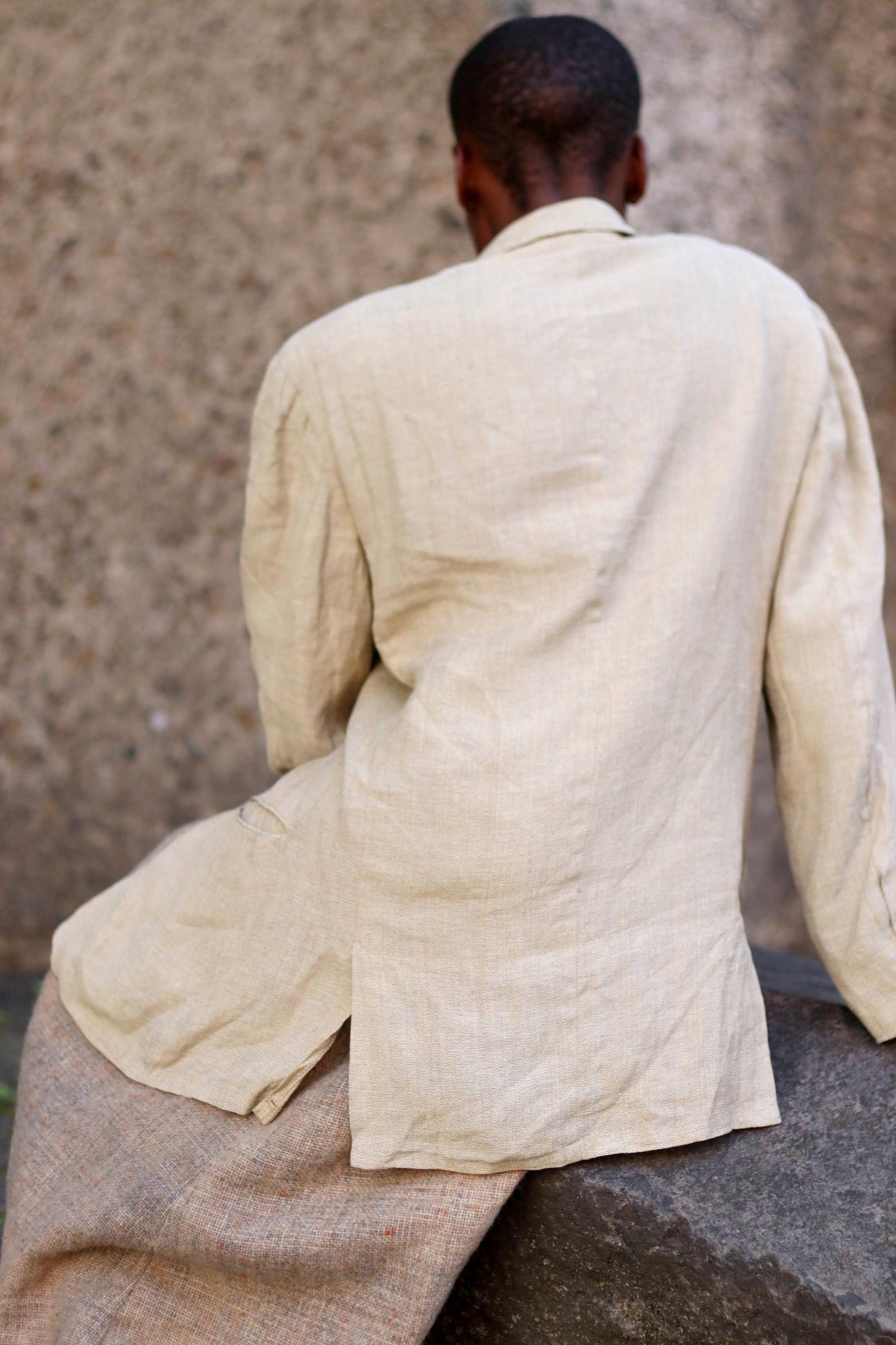 Female Model of African Heritage in a vintage Hugo Boss beige linen blazer from the 90s paired with a brown skirt photographed  against a textured stone background. She sits with her face away from the camera