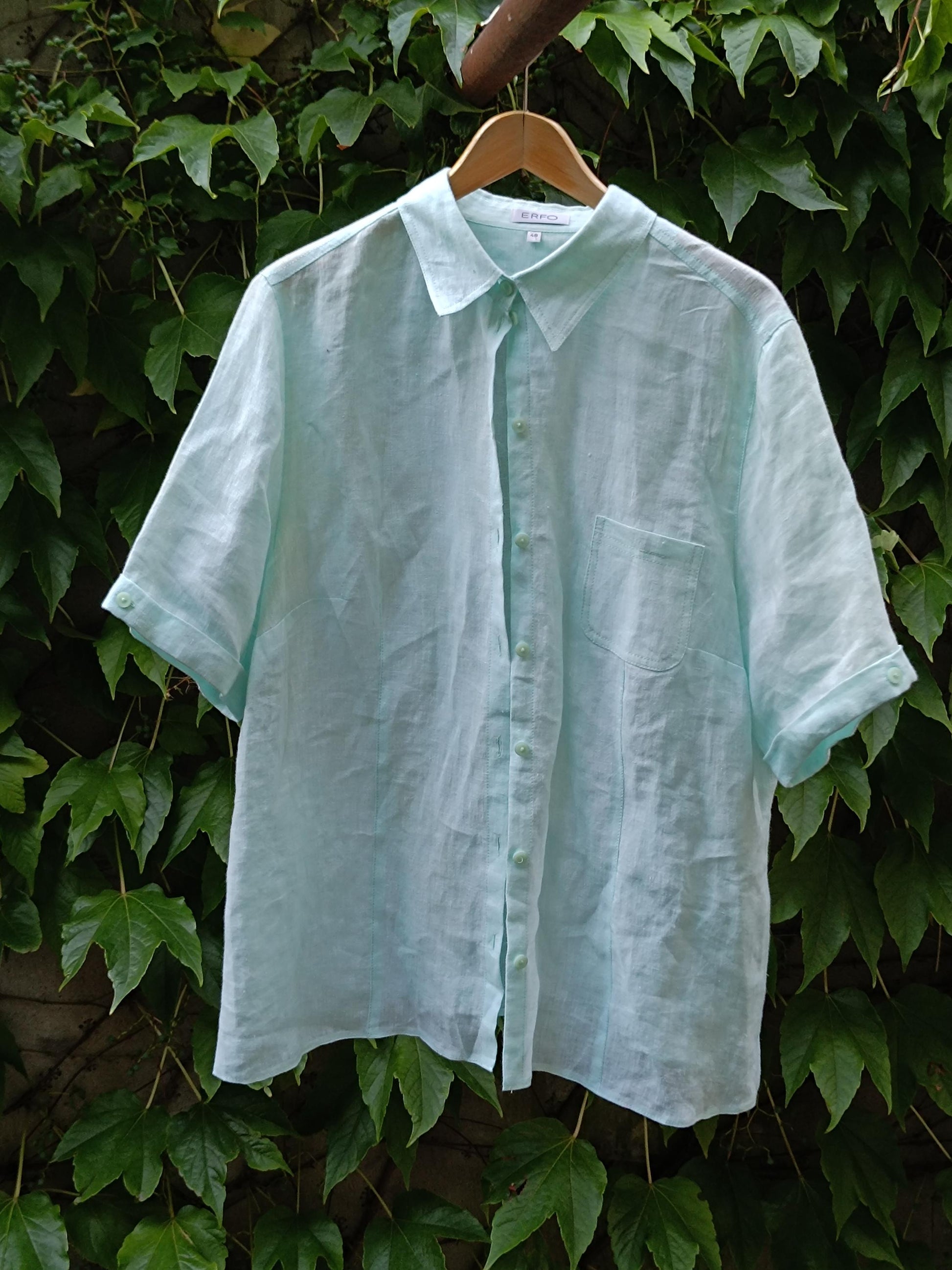 A 90s vintage light blue, relaxed-fit linen shirt with short-sleeved shirt is photographed hanging from a hanger on a clothing line against a background of green leaves. The minimalist shirt is perfect for summer