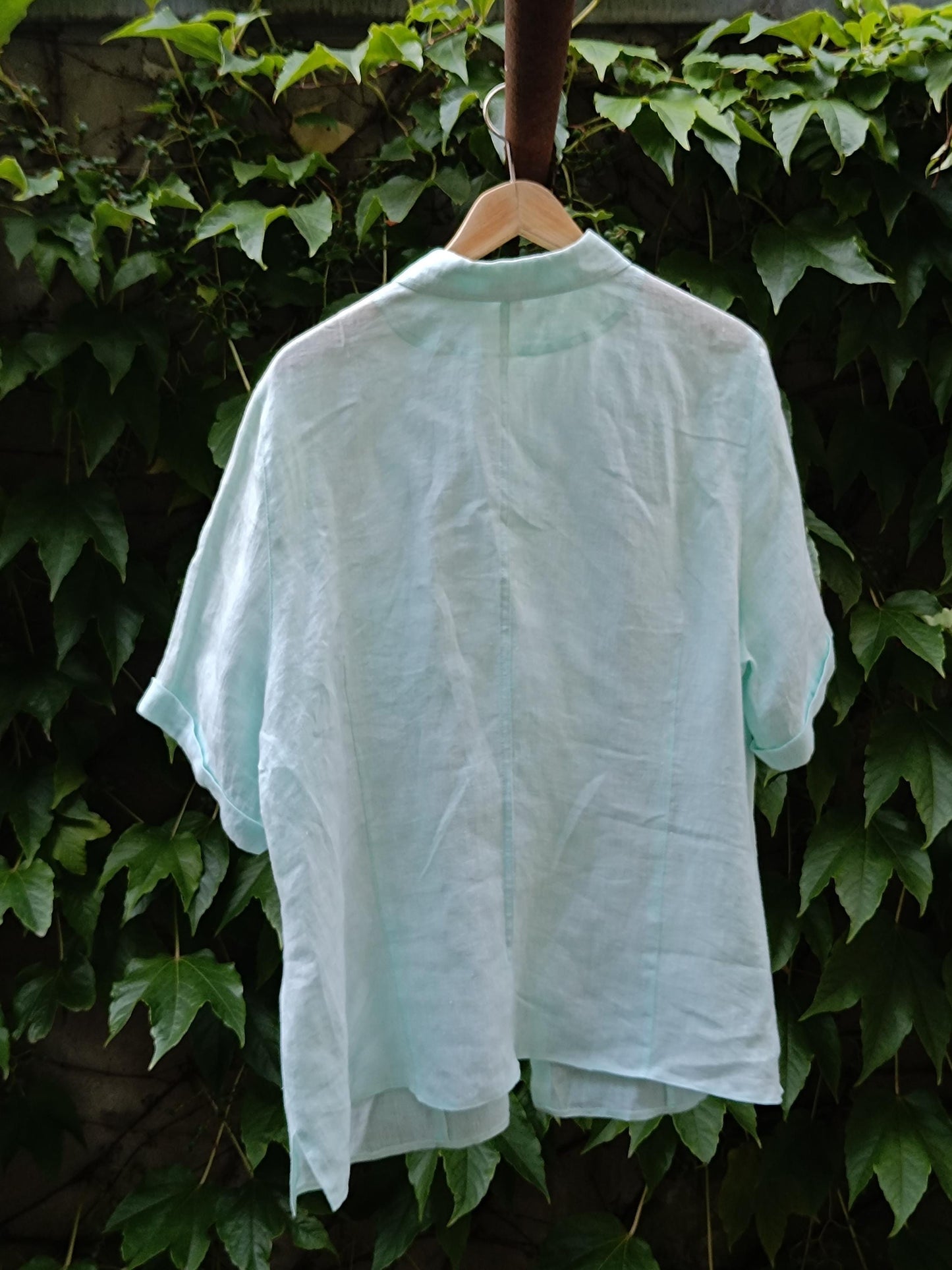 A back view of a 90s vintage light blue, relaxed-fit linen shirt. The short-sleeved shirt is photographed hanging from a hanger on a clothing line against a background of green leaves.