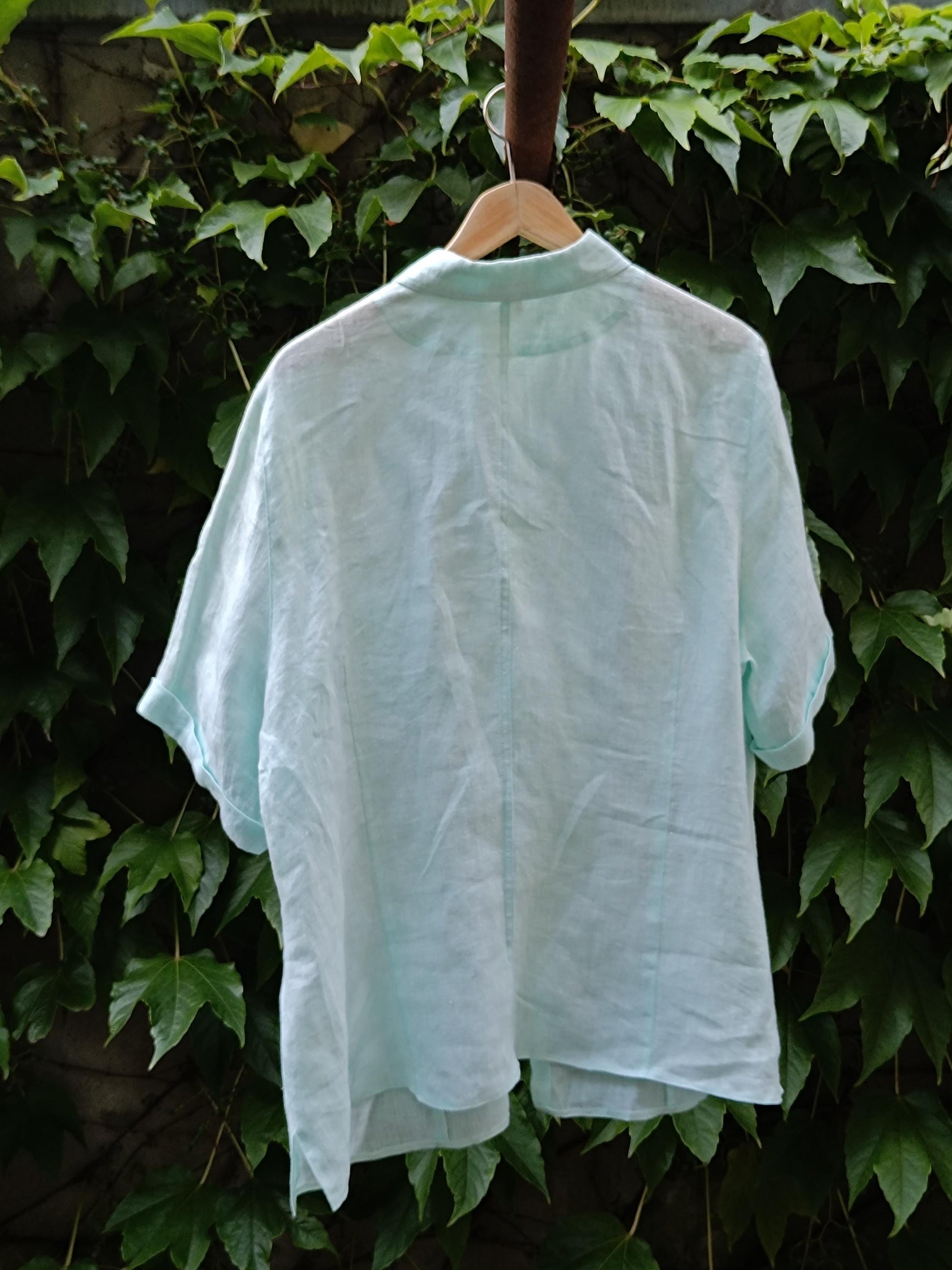 A back view of a 90s vintage light blue, relaxed-fit linen shirt. The short-sleeved shirt is photographed hanging from a hanger on a clothing line against a background of green leaves.