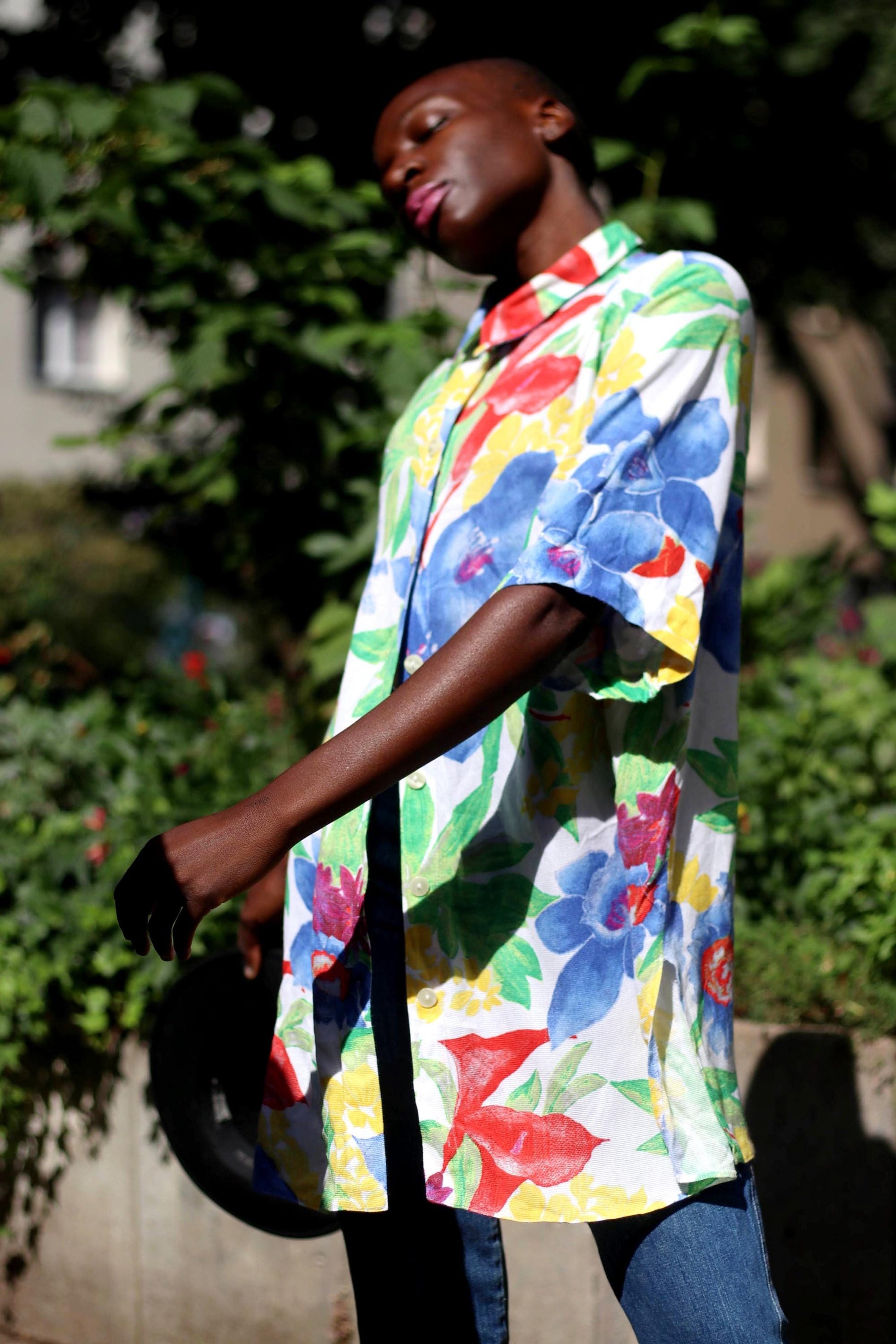Fashionable model of African heritage poses in a colorful vintage floral print shirt and jeans combo. She stands against a green leafy background with her eyes are closed