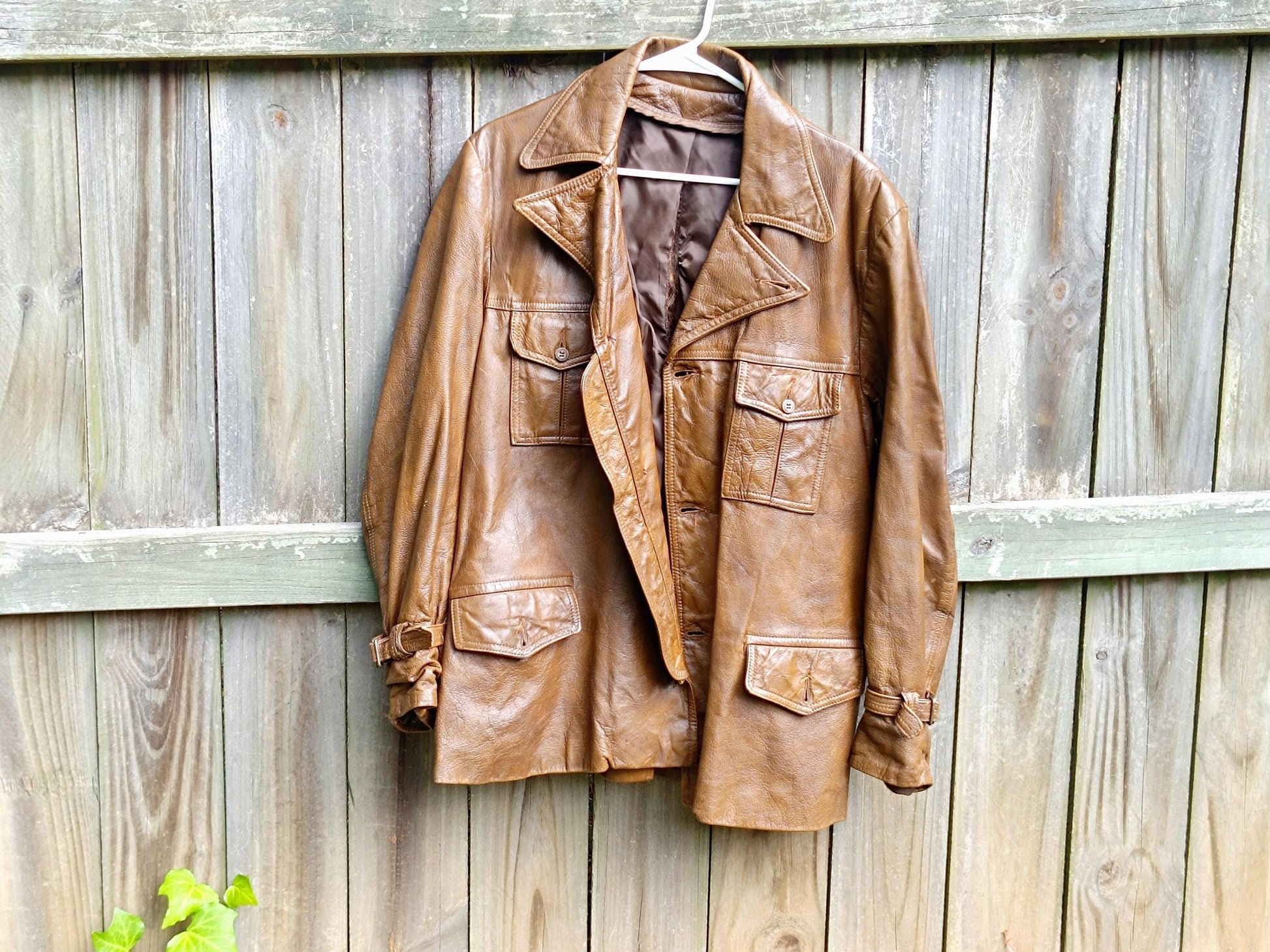 A 90s Vintage Leather Jacket in brown is photographed hanging on a hanger Infront of a brown rustic wooden fence. The image highlights the huge and minimalist cool silhouette