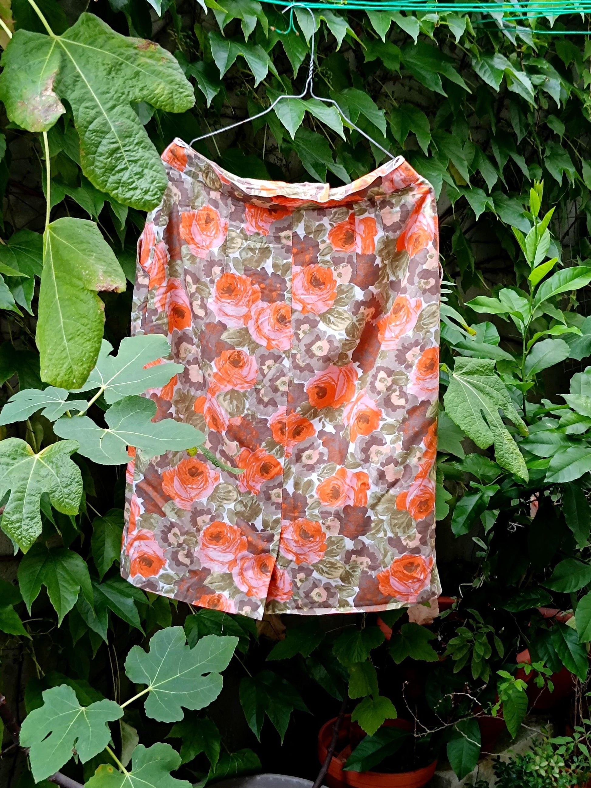 An 80s  Orange Floral Skirt photographed hanging from a hanger with green plants in the background. The image showcases the bold print on the vontage knee-length Skirt
