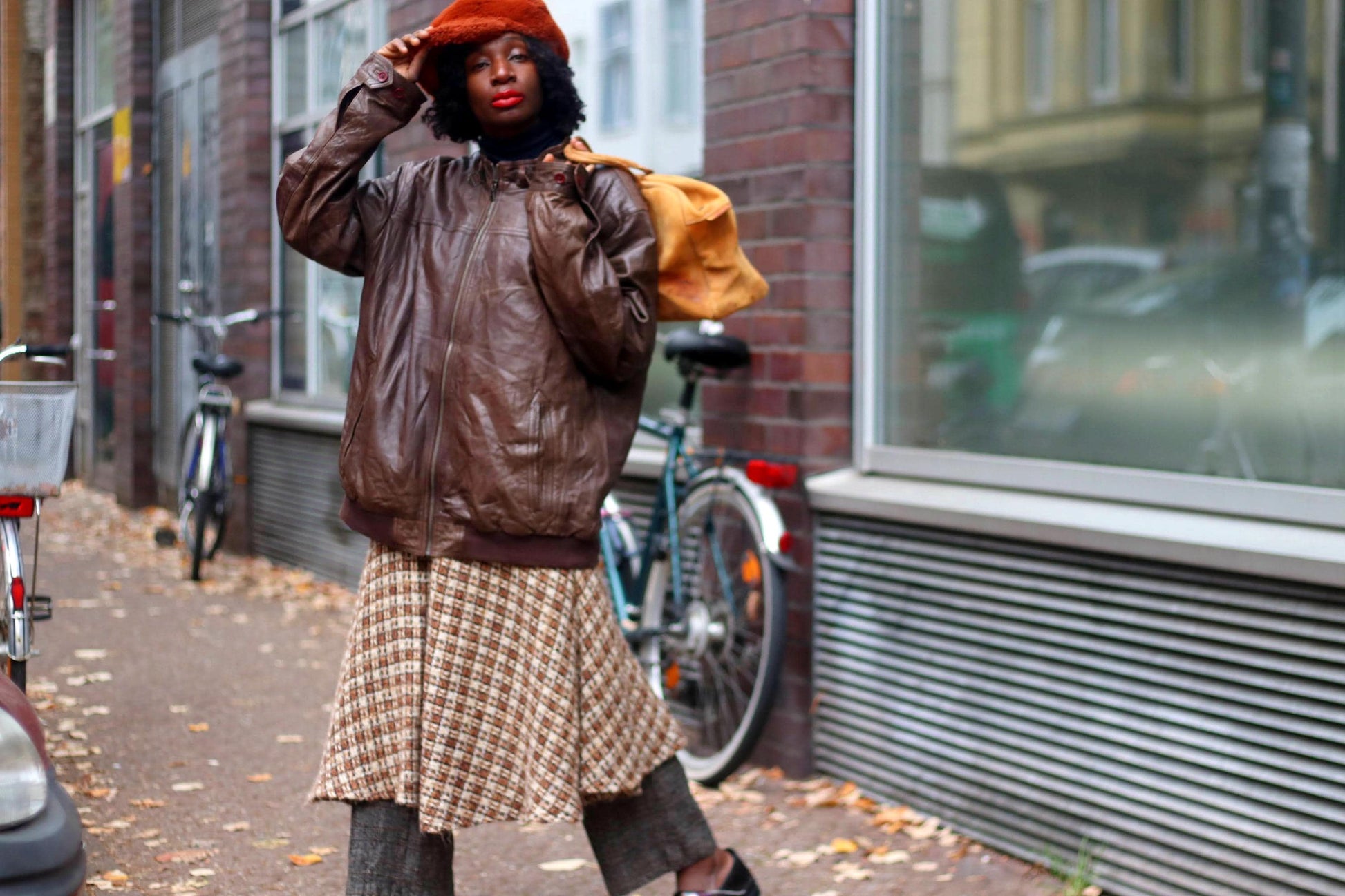 Stylish woman in vintage brown leather jacket and plaid skirt posing on urban street with bicycles, showcasing chic fall fashion.