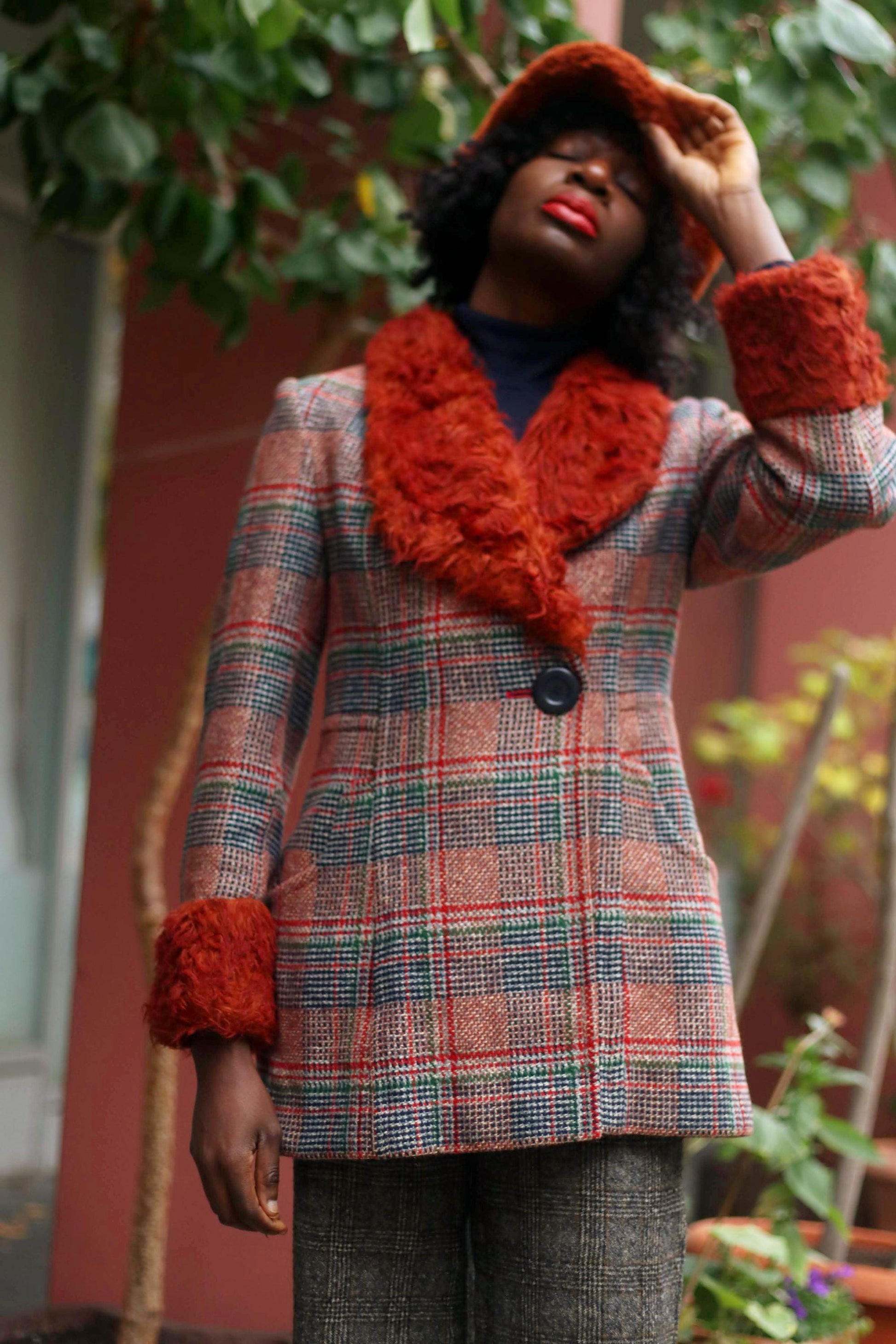 Person wearing a vintage Gil Bret plaid coat with a burnt-orange faux fur collar and cuffs, holding the brim of a hat. The coat has a single black button and deep front pockets. Background includes a pink wall & green plants, creating an autumn vibe