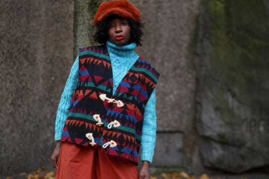 Fashionable individual wearing a vintage-inspired vest with geometric patterns, paired with a blue cable-knit sweater, a terracotta beret, and orange trousers. Styled outdoors with a natural, textured background, highlighting fall fashion trends
