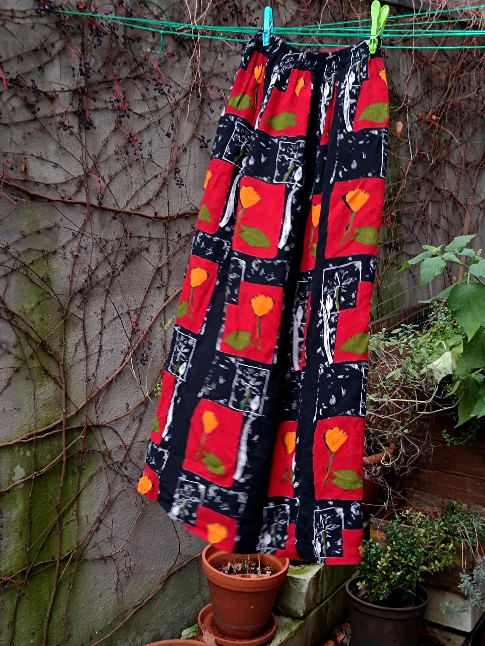 A still life image of a  vintage red skirt with floral pattern with red blocks of yellow flowers and leafy green accents. The retro 90s maxi skirt is photographed hanging from a clothing line in a garden with a weathered wall with twines as background and green leaves at the back. The skirt has a side slit