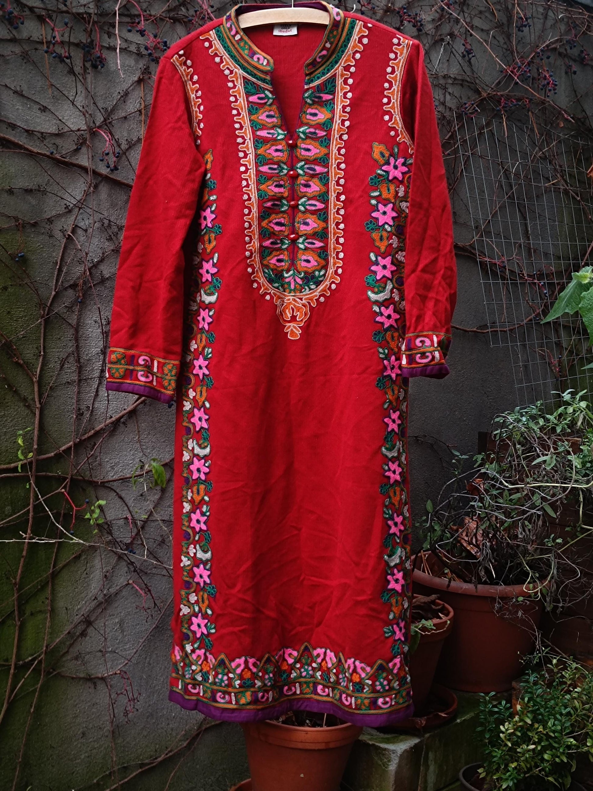 Bold vintage 70s red dress with intricate embroidery, bohemian fashion, long sleeve dress, hanging against a weathered wall partially covered in vines, rustic charm.