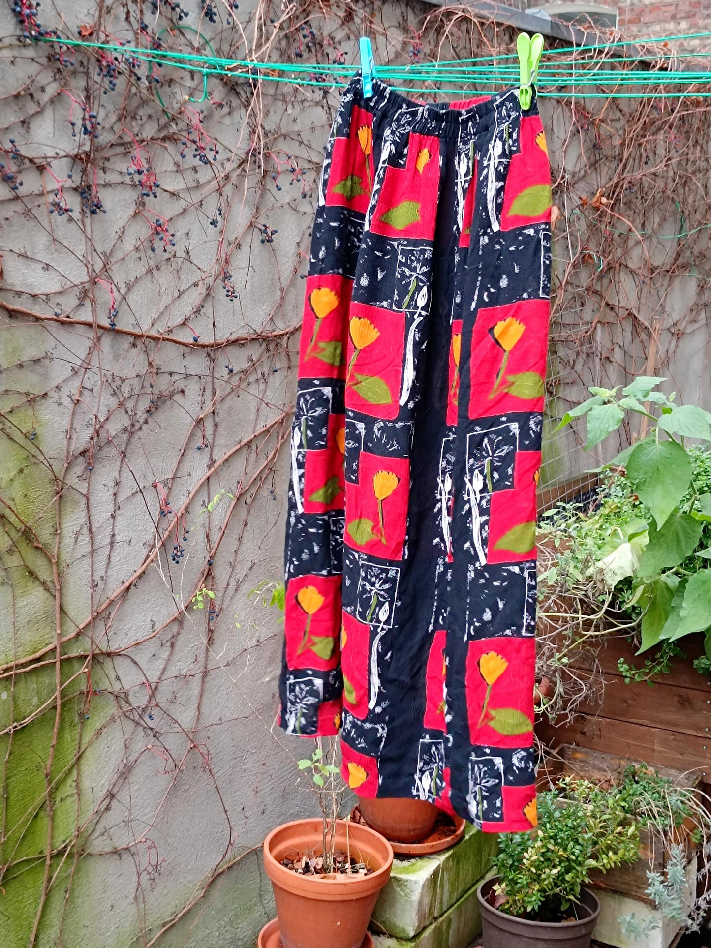 A vintage red skirt with floral pattern showing red blocks with yellow flowers and leafy green accents is photographed hanging from a clothing line in a garden with a weathered wall with twines as background and green leaves at the back. emphasizing the retro boho print and summer vibe.