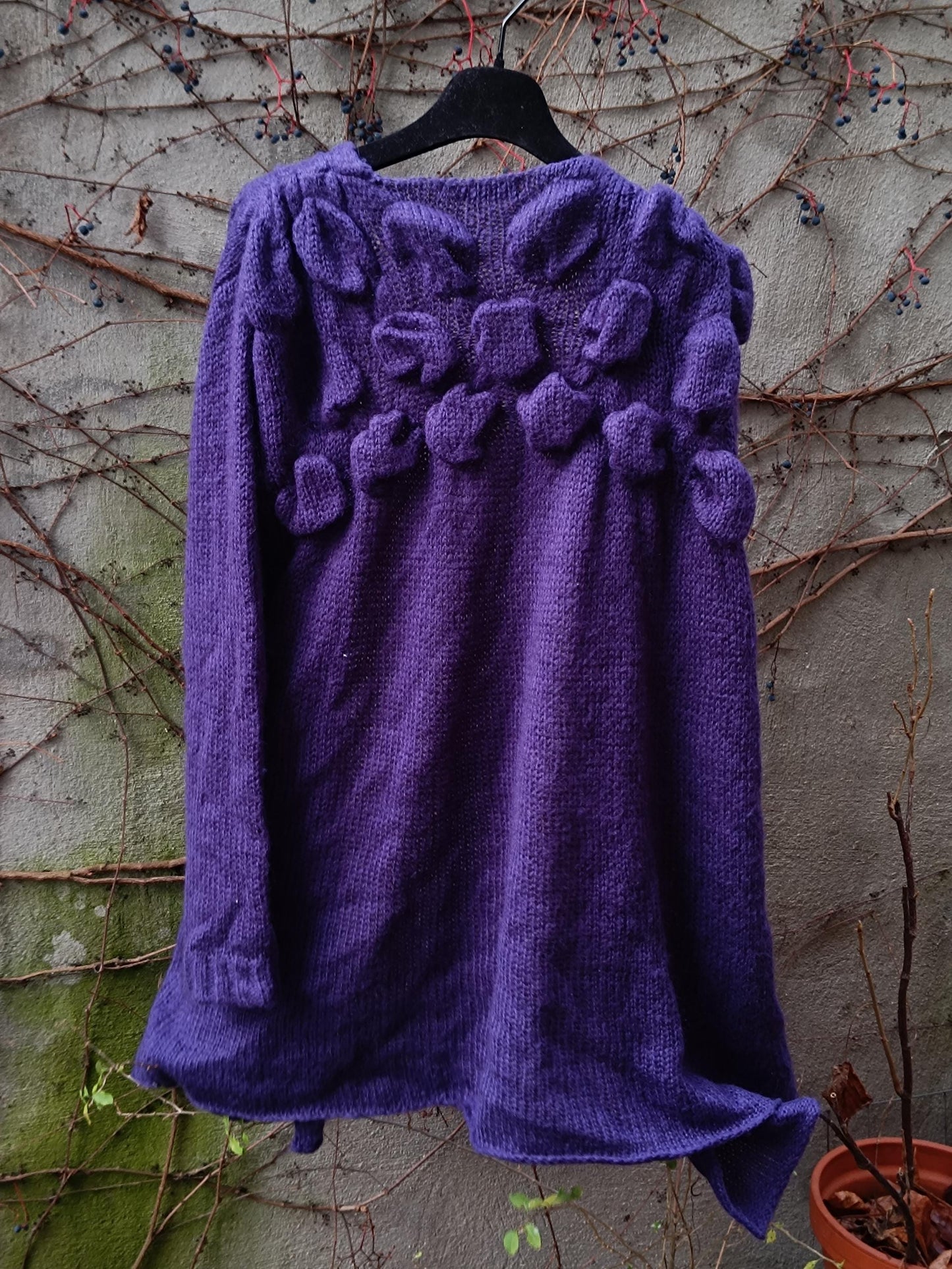 Back view of a purple floral cardigan by Mais il est où le Soleil, photographed against a weathered grey wall with climbing plants