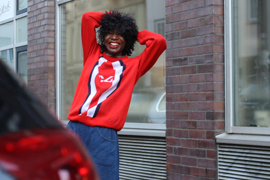 Woman smiling in a vibrant red and white vintage striped sweater, standing next to a car  in an urban setting, showcasing street fashion and casual style.