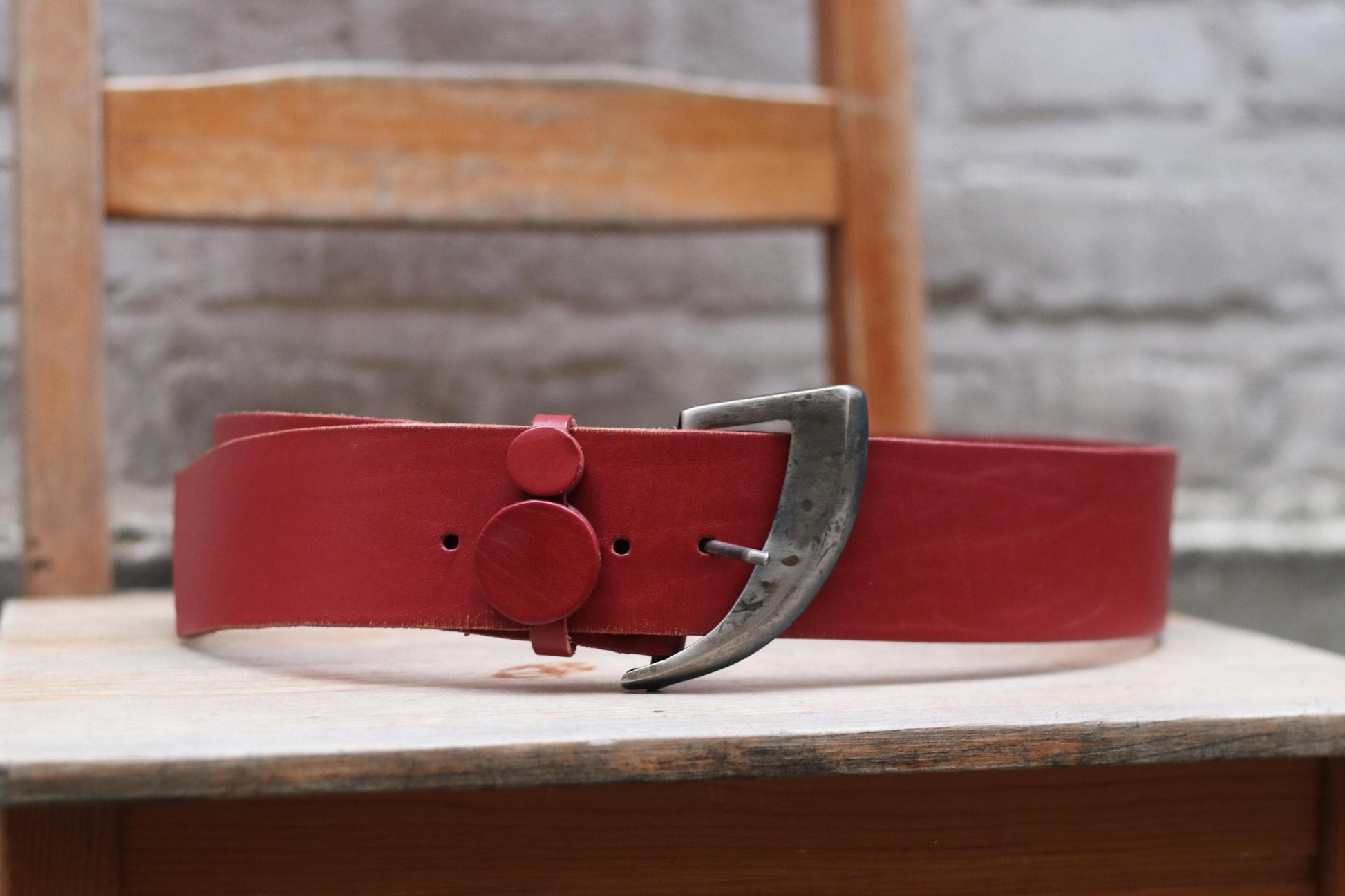 Vintage red leather belt with a rustic silver buckle displayed on a wooden chair against a brick wall background