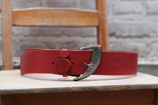 Vintage red leather belt with a rustic silver buckle displayed on a wooden chair against a brick wall background