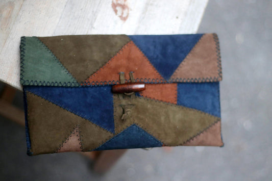 A vintage clutch made of patchwork suede in various earthy tones, including navy, brown, green, and terracotta. The 90s handmade clutch has a wooden toggle closure and is resting on a light-coloured surface photographed with a bird's eye view.