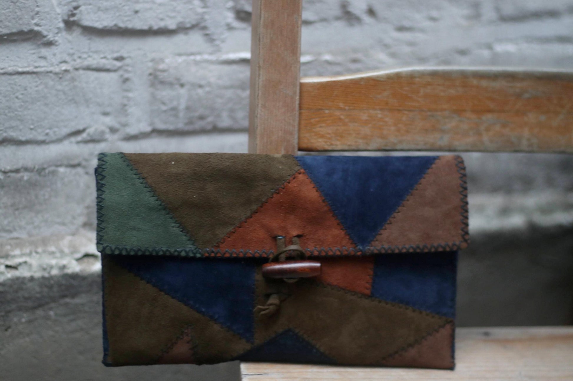 A vintage clutch made of patchwork suede in various earthy tones, including navy, brown, green, and terracotta. The clutch has a wooden toggle closure and is resting on a light-coloured surface.