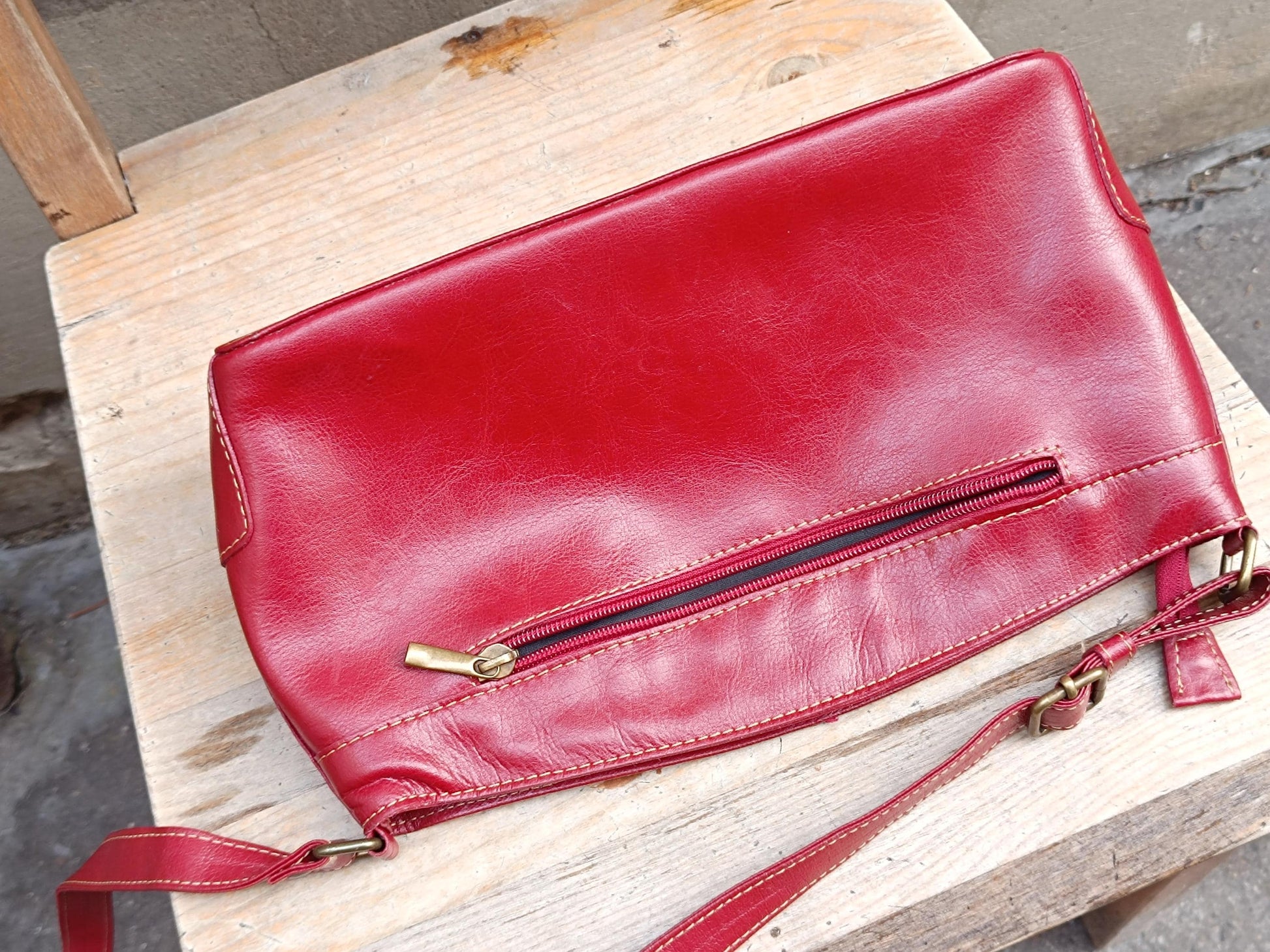 Vintage 90s red leather shoulder bag with a classic design is captured on a simple brown wooden chair. The image emphasizes the back of the back, showcasing its soft leather and functional details like the zipper and adjustable strap.