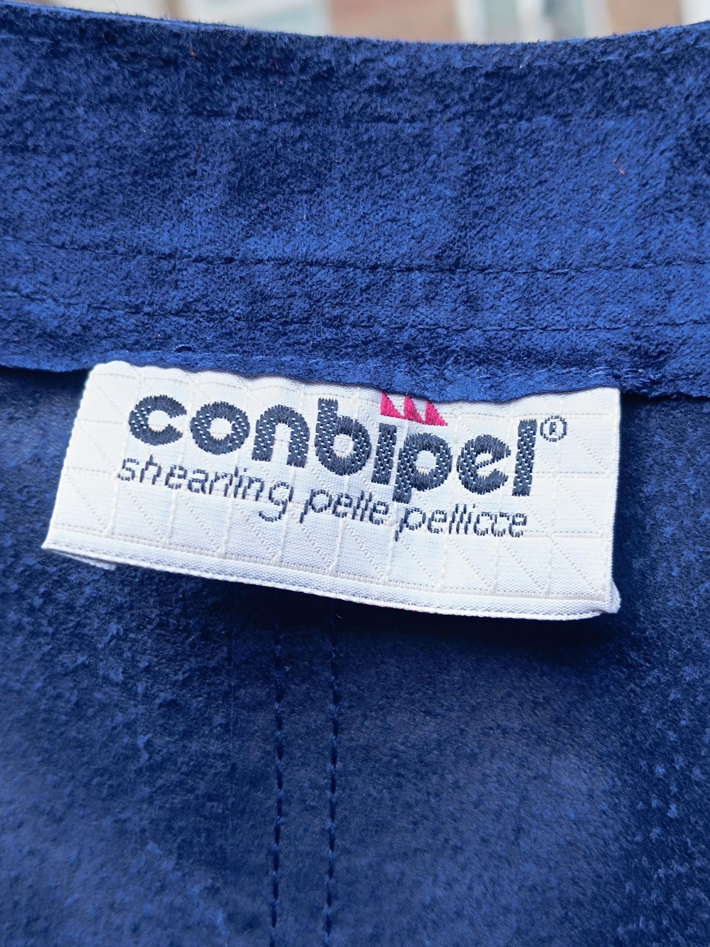 A close up image of a white label with the text &#39;conbipel® shearing pelle pellicce&#39; in black text on a blue suede fabric background.