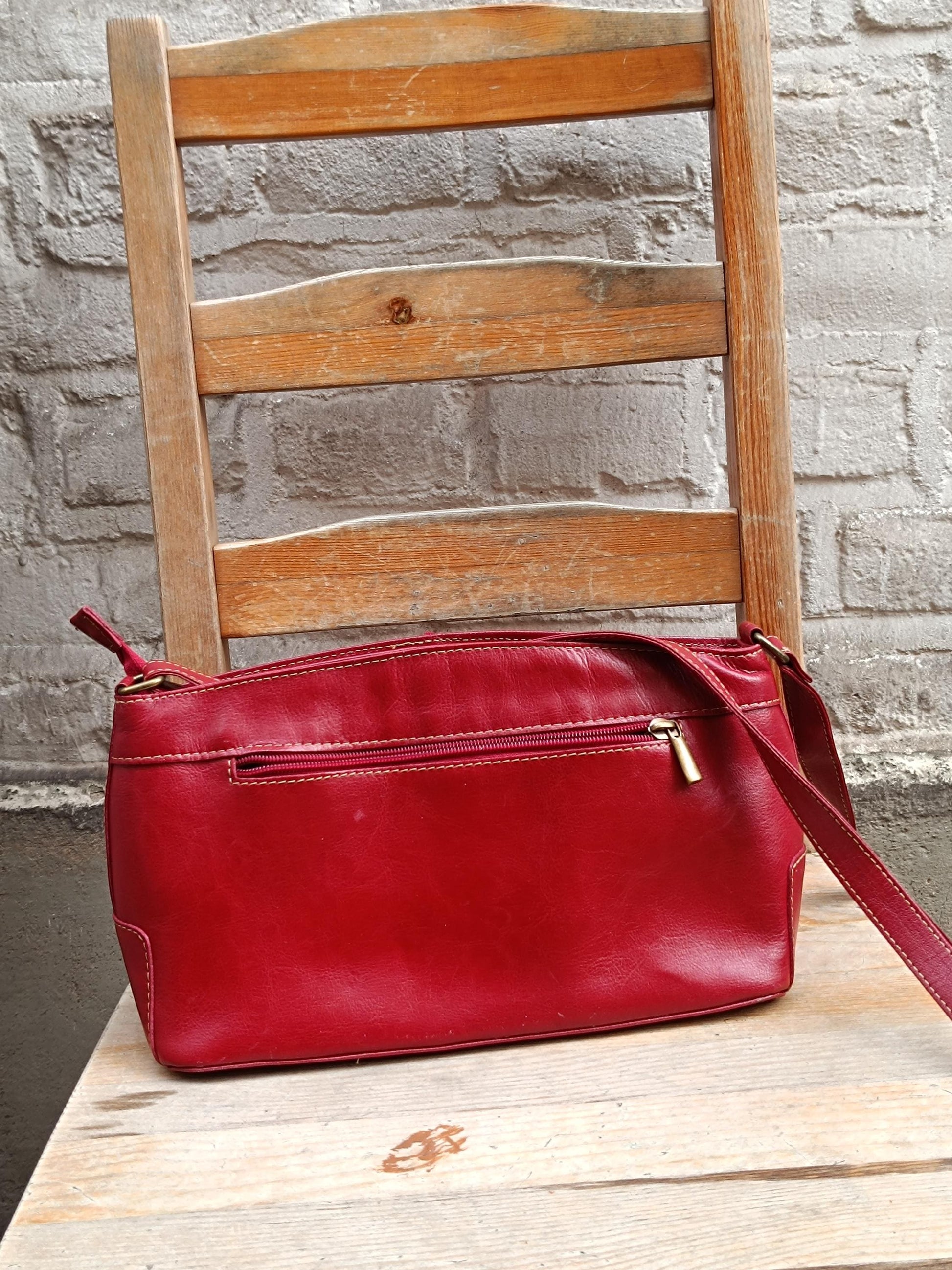 Vintage red leather shoulder bag  from the 90s with a single adjustable strap and silver hardware is displayed on a light wooden chair against a textured gray wall. The bag has a visible zipper closure.