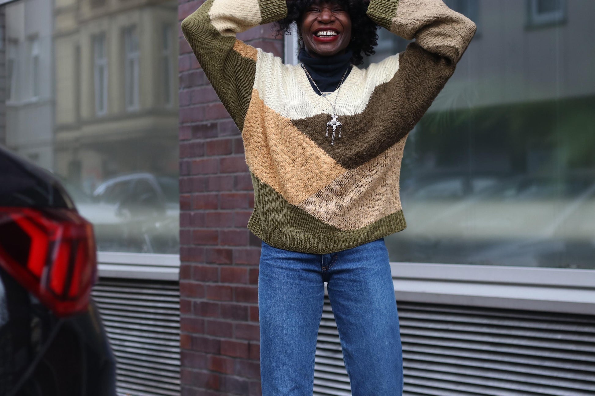 Smiling female model wearing a colorful  70s vintage handmade patchwork sweater with brown,  green and orange hues paired with blue jeans, playfully posing on a city street.