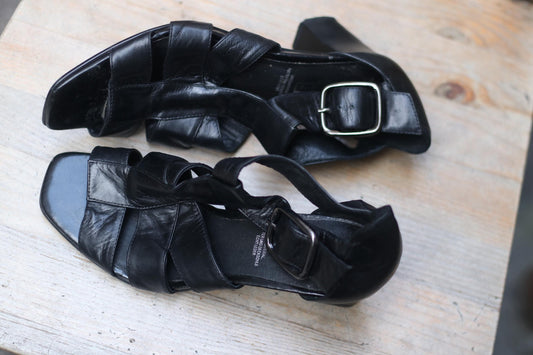 A pair of vintage 5th Avenue black leather sandals featuring a woven strappy design, square toe, and chunky block heel. The 90s summer shoe is photographed on a wooden surface.