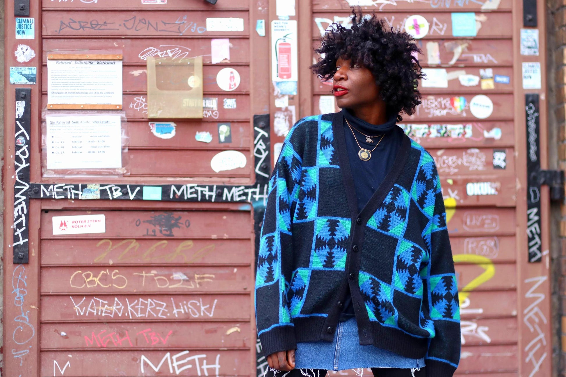 A woman poses in front of an urban, graffiti-covered backdrop wearing a vintage Ackermann oversized cardigan with a bold blue & black geometric print. Paired with a denim mini skirt,and gold jewelry, the look is effortlessly chic, retro, and modern
