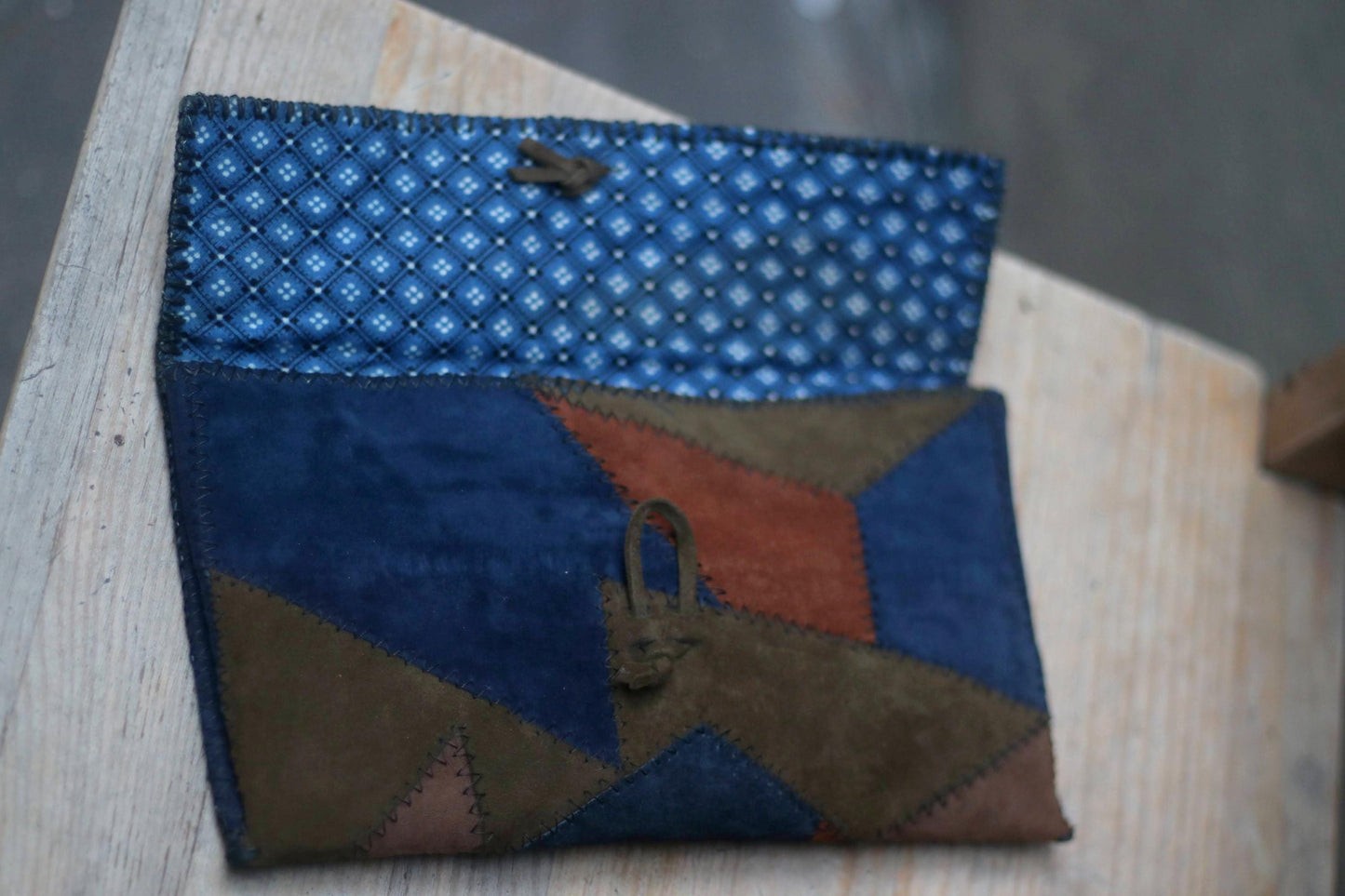 A vintage clutch made of patchwork suede in various earthy tones, including navy, brown, green, and terracotta. The clutch has a wooden toggle closure and is resting on a light-coloured surface. the inside lining is blue